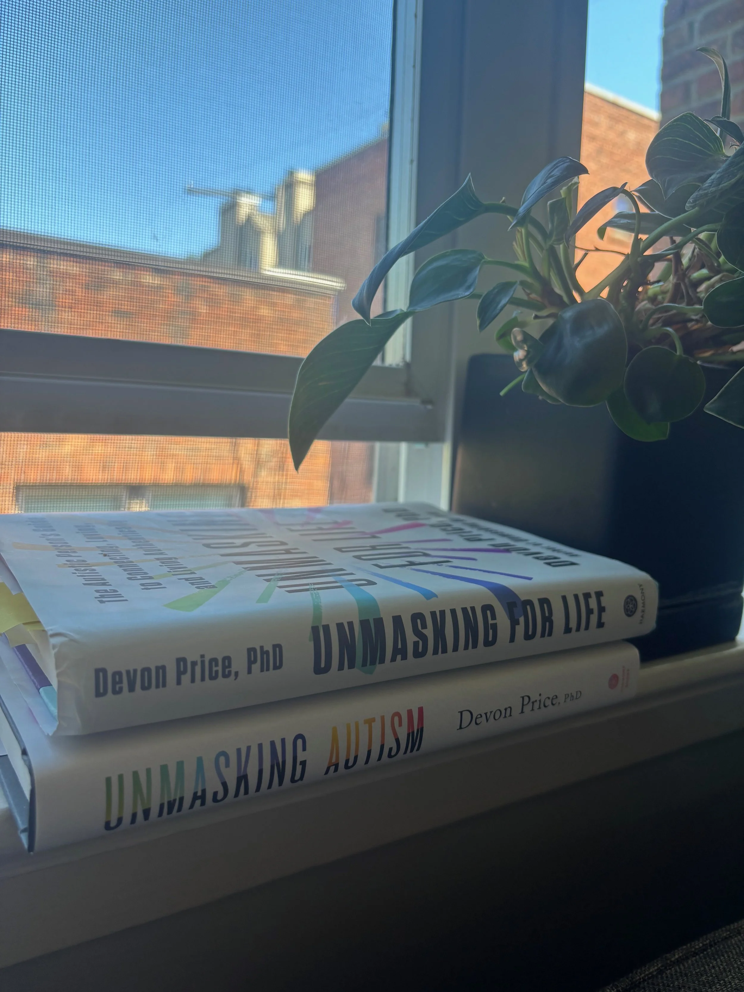 Two hardcover books sit a windowsill next to a plant overlooking a sunny say and brick buildings outside