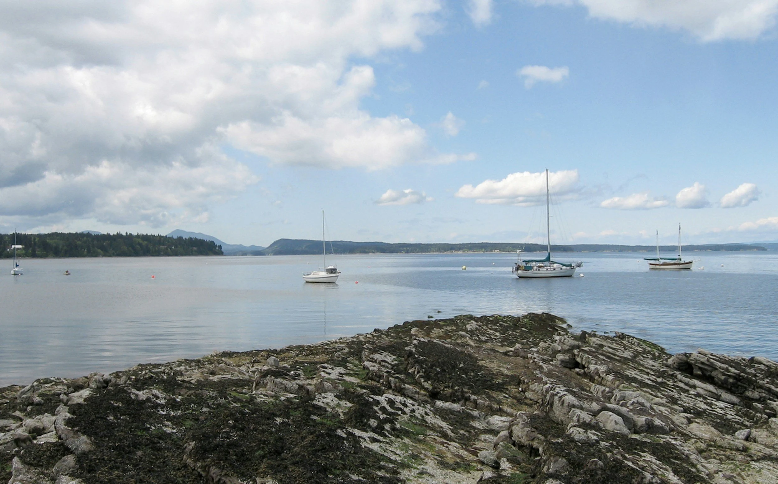 A peaceful scene of sailboats on calm water near rocky shoreline with distant hills under a partly cloudy sky.