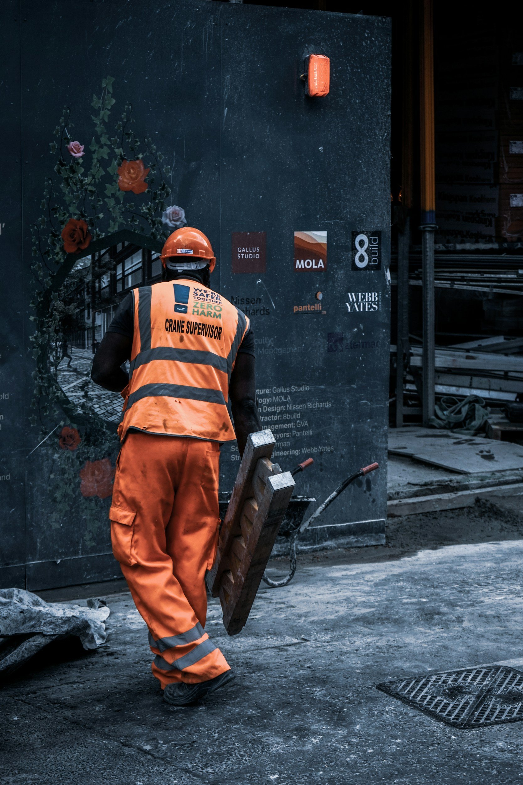 A construction worker wearing an orange vest and helmet, with 'CRANE SUPERVISOR' written on the back, walking on a damaged street holding wooden planks. There are various company logos and text on the wall behind him, and construction materials are visible.