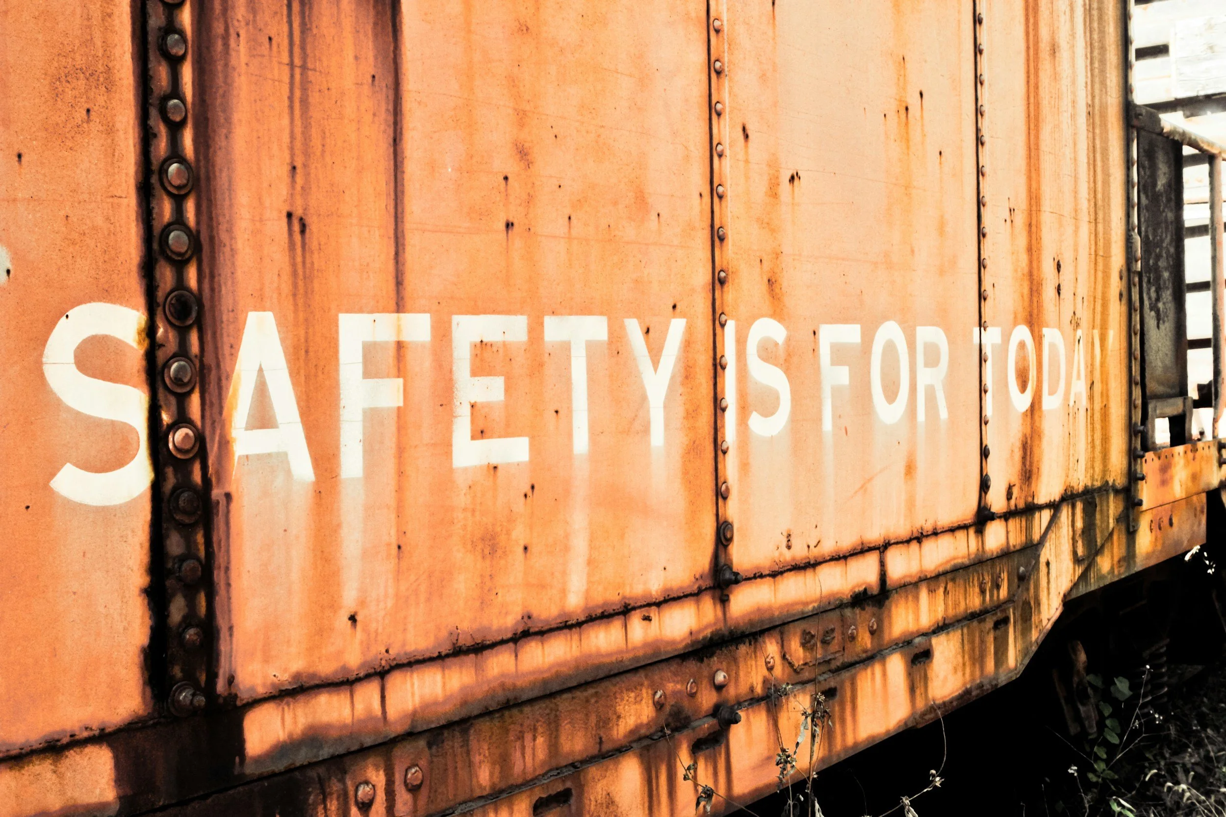 Close-up of an old, rusty orange train car with white partially visible text that reads 'SAFETY IS FOR TODAY' and visible rivets.