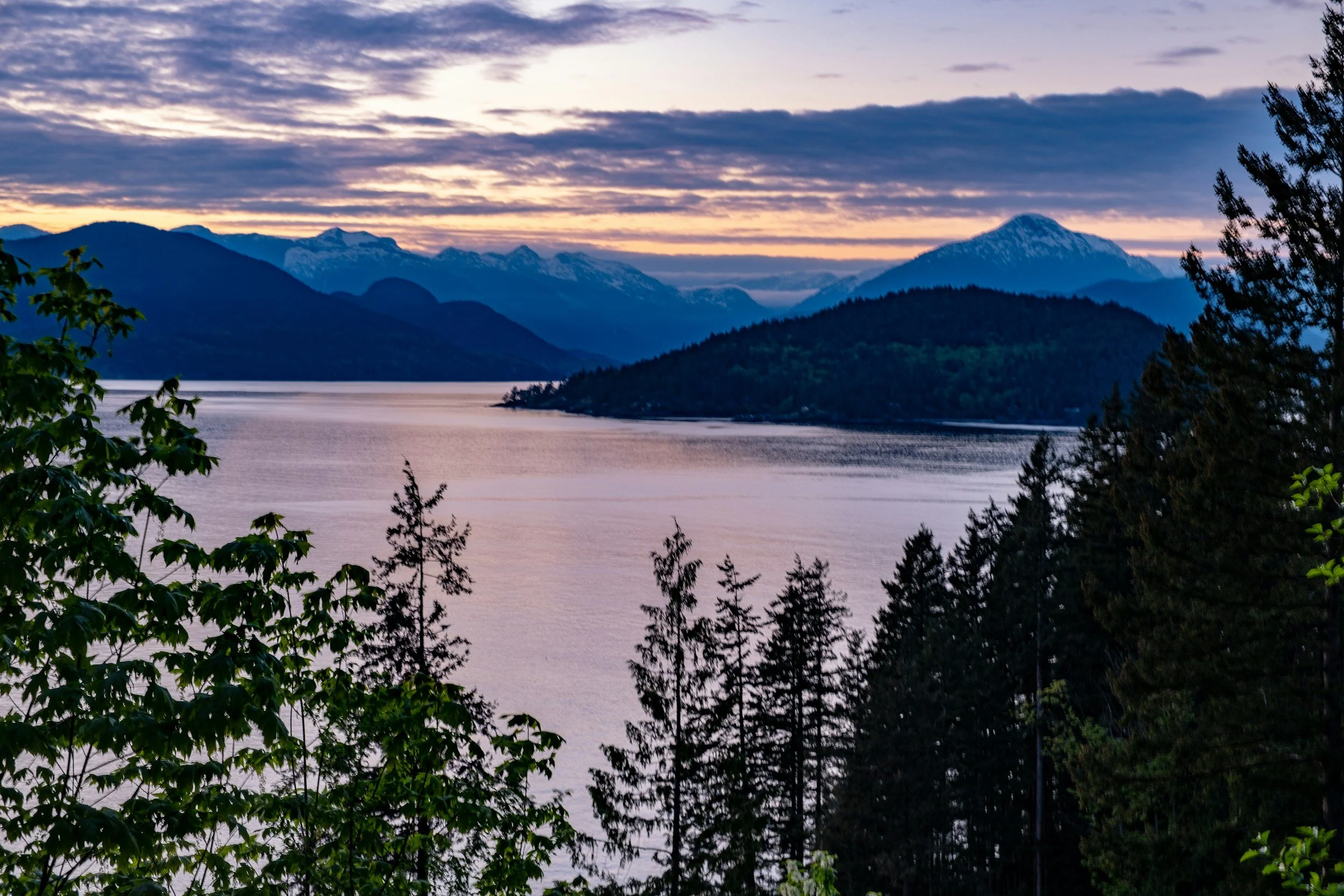 A scenic view of a lake with a mountain range in the background, trees in the foreground, and a colorful sky at sunset.