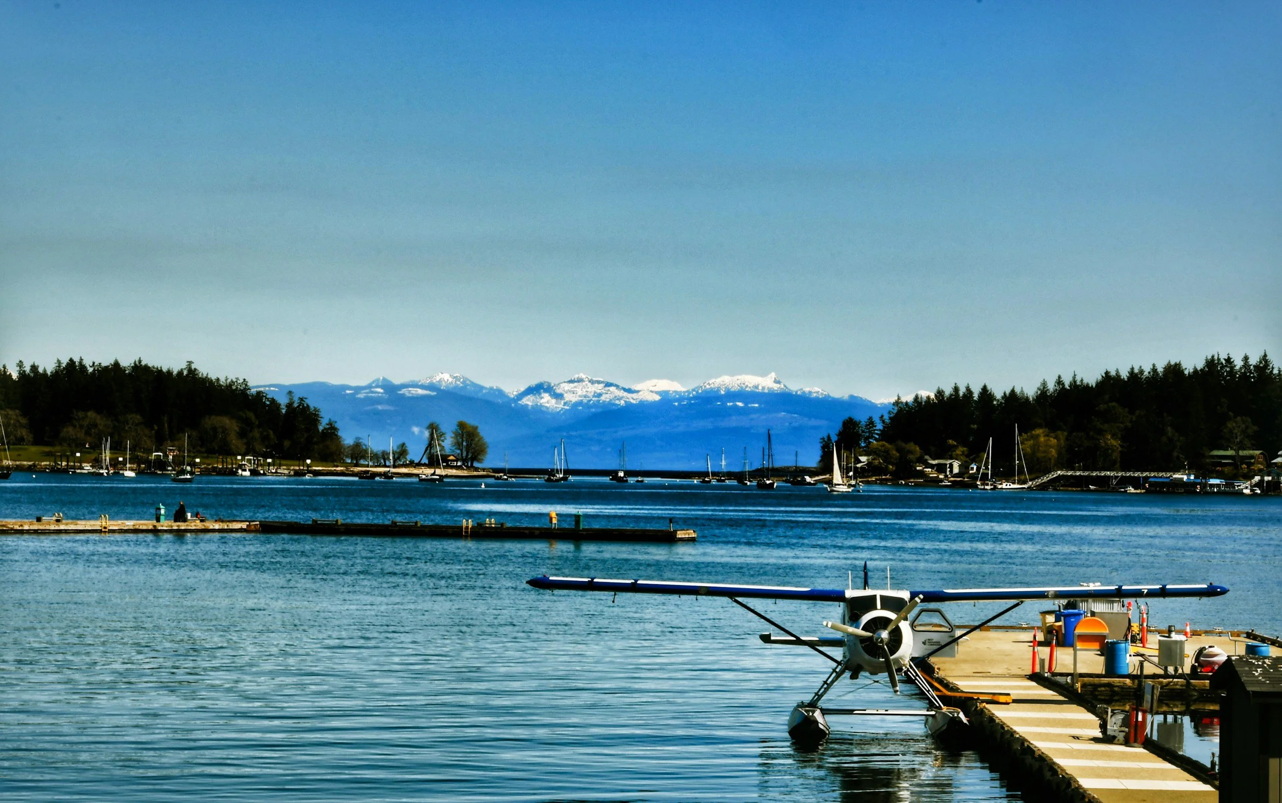 A waterfront scene with boats anchored in a calm bay, a pier with a small airplane, and snow-capped mountains in the distance under a clear blue sky.