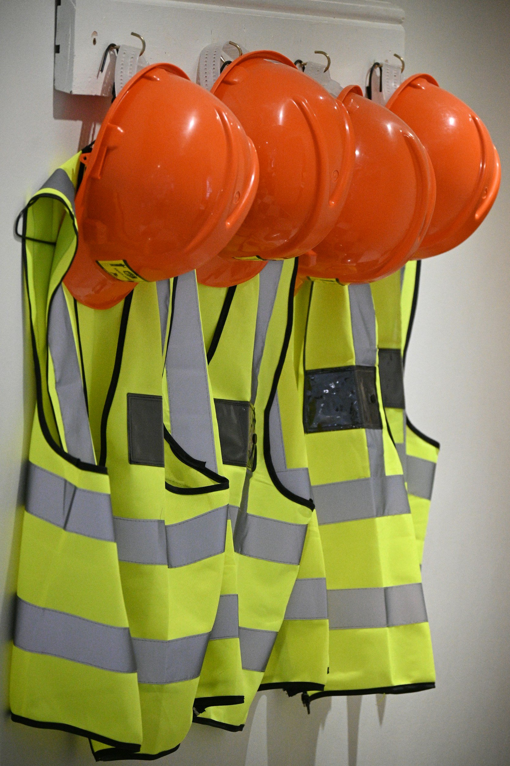 A row of four orange safety helmets and four high-visibility yellow safety vests hanging on hooks against a plain wall.