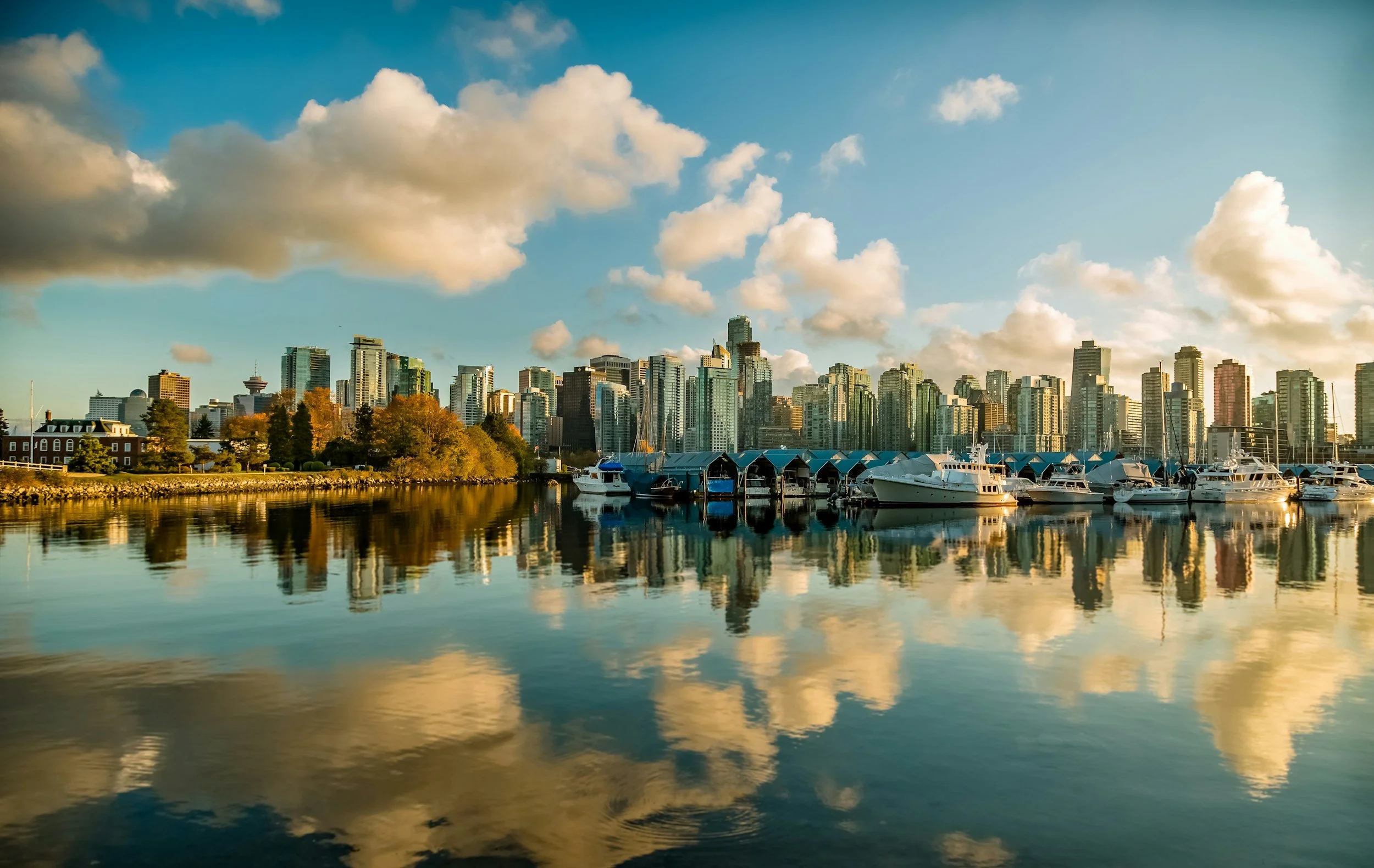 City skyline with tall buildings overlooking a marina with boats, reflected in calm water, under a partly cloudy sky during daylight.