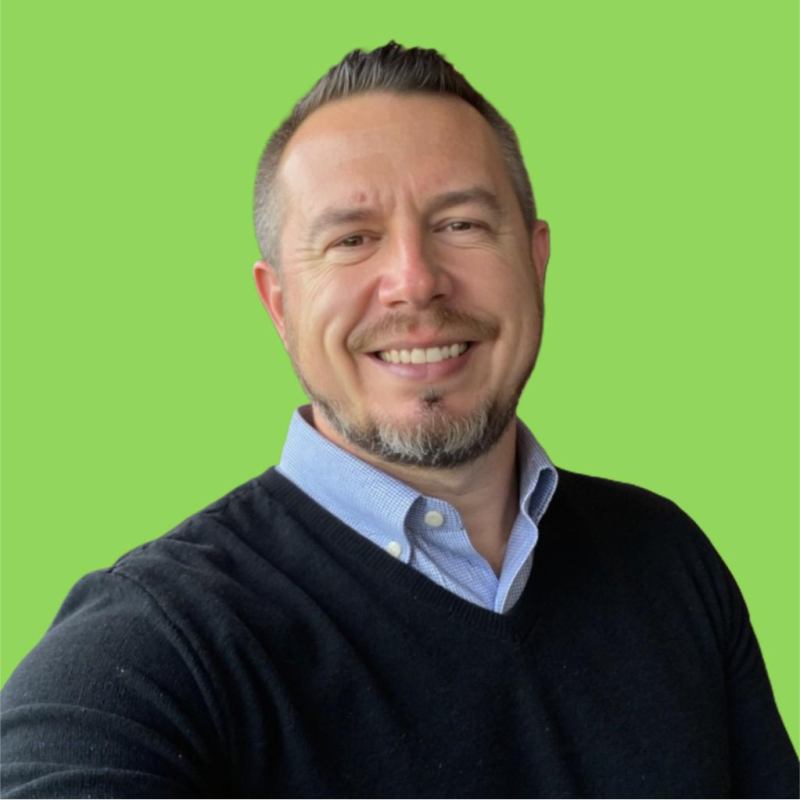 A smiling man with short brown hair, facial hair, wearing a light blue collared shirt and a black sweater, standing against a bright green background.