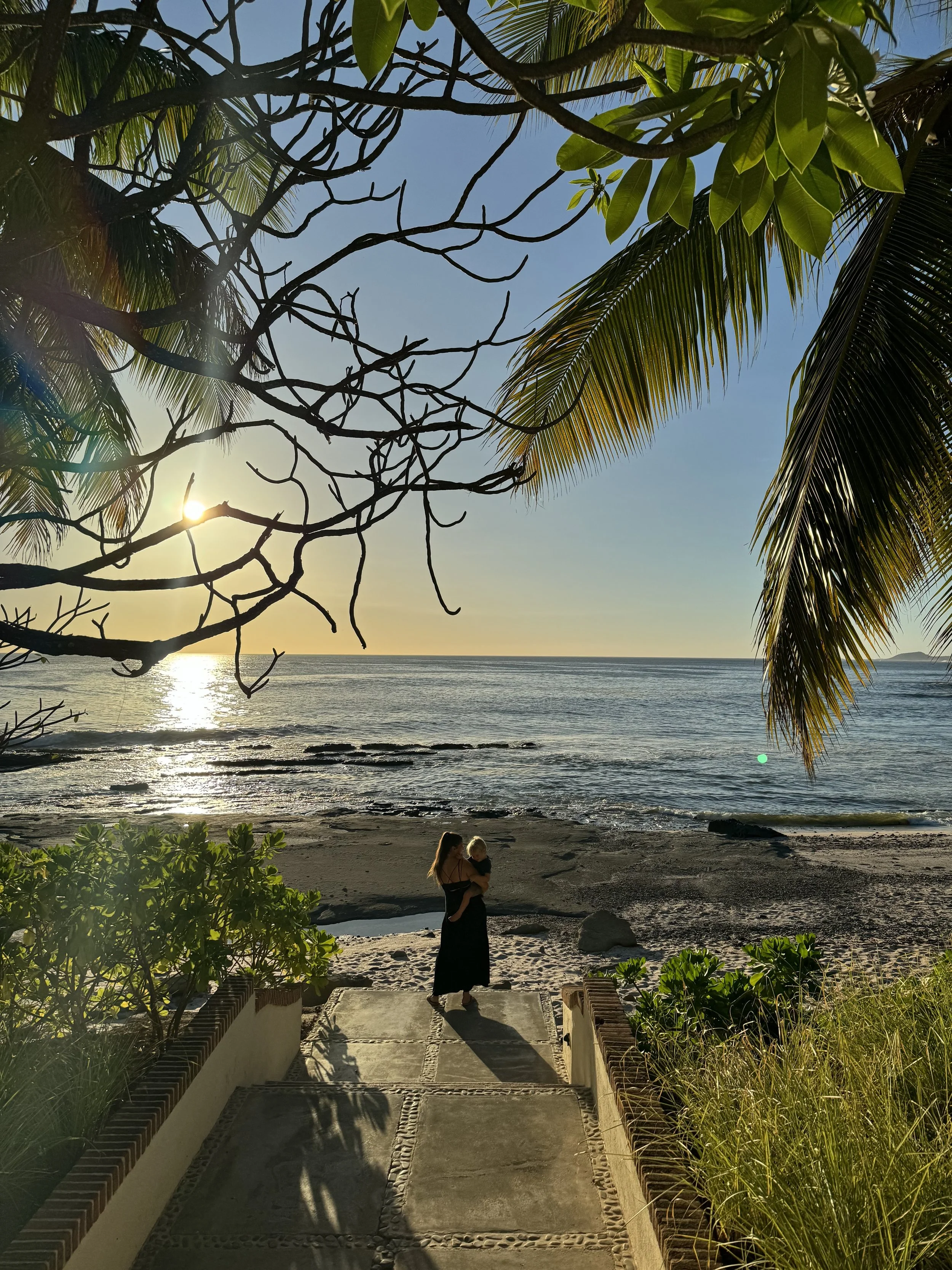 Woman holding a toddler on the beach overlooking the ocean and surrounded by lush greenery
