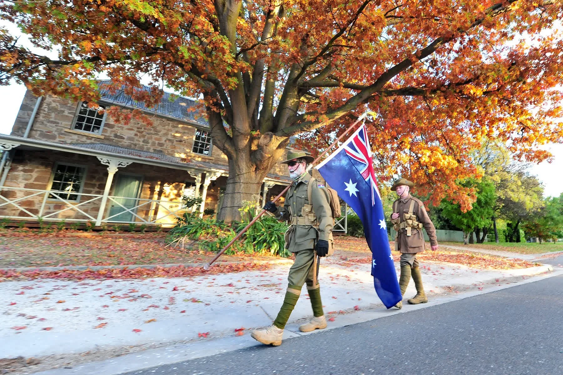 Two men dressed in World War II military uniforms walking along a tree-lined street during autumn, with the unknown veteran holding an Australian flag and the other walking beside him heading from Sydney to Canberra for Anzac Day..