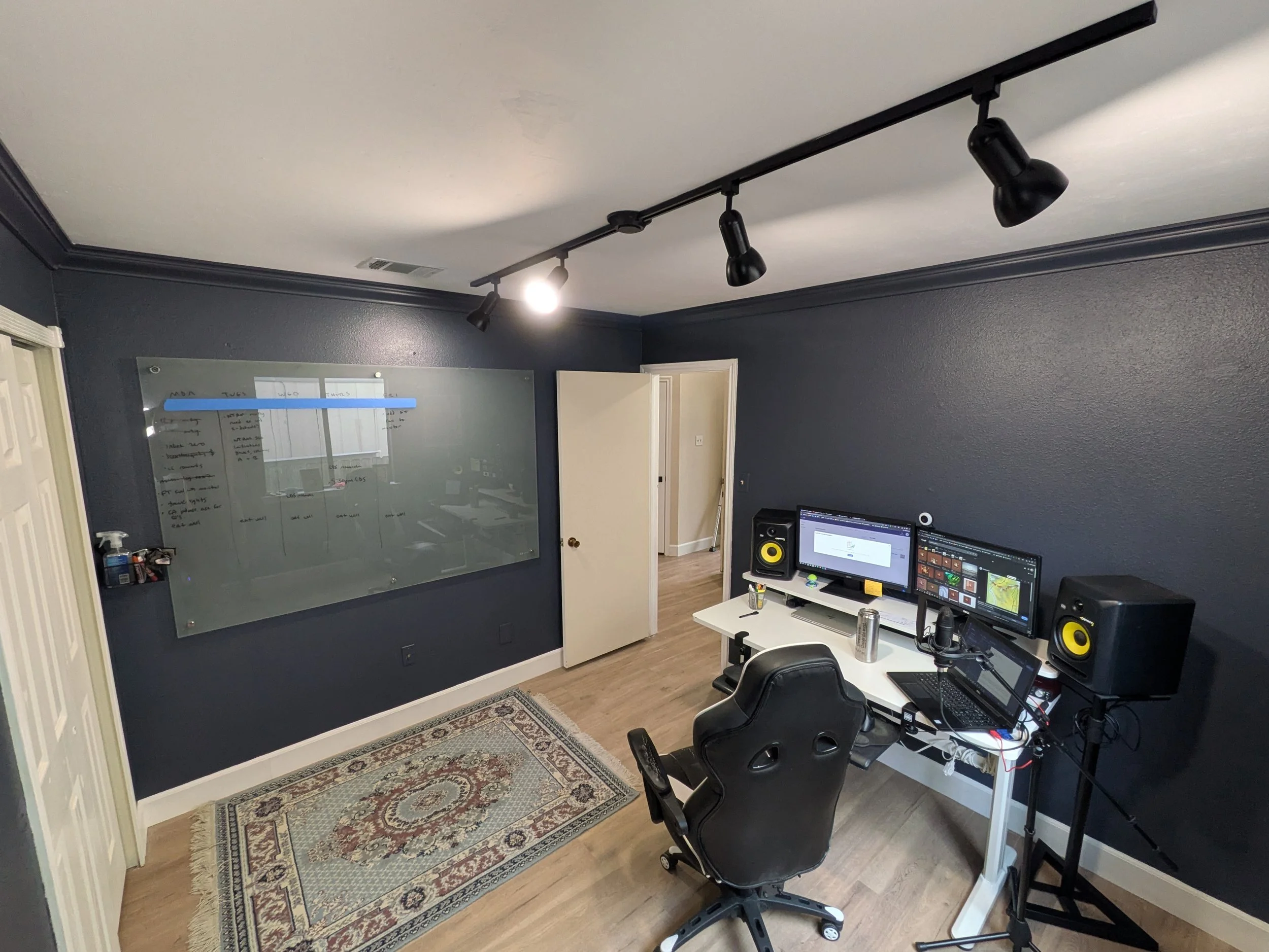 a home office - dark blue walls, a light blue rug, a large dry erase board and a desk setup with 3 monitors and large studio speakers