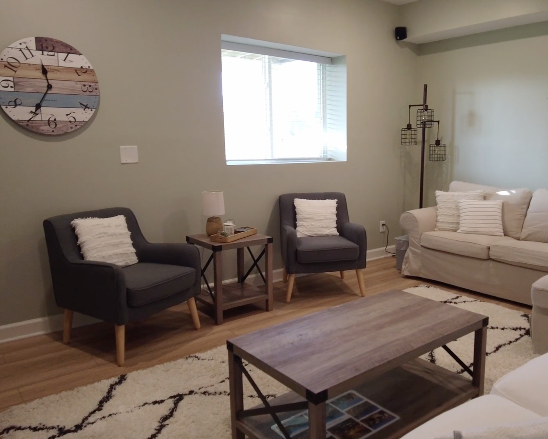 Living room with a beige sofa, two gray armchairs, a wooden coffee table, and a small side table with a lamp. There is a wall clock on the wall and a window letting in natural light.