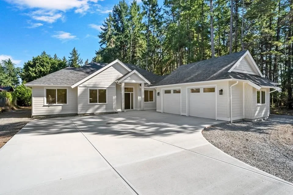 A white modern house with a three-car garage and a spacious concrete driveway, surrounded by tall trees under a blue sky.