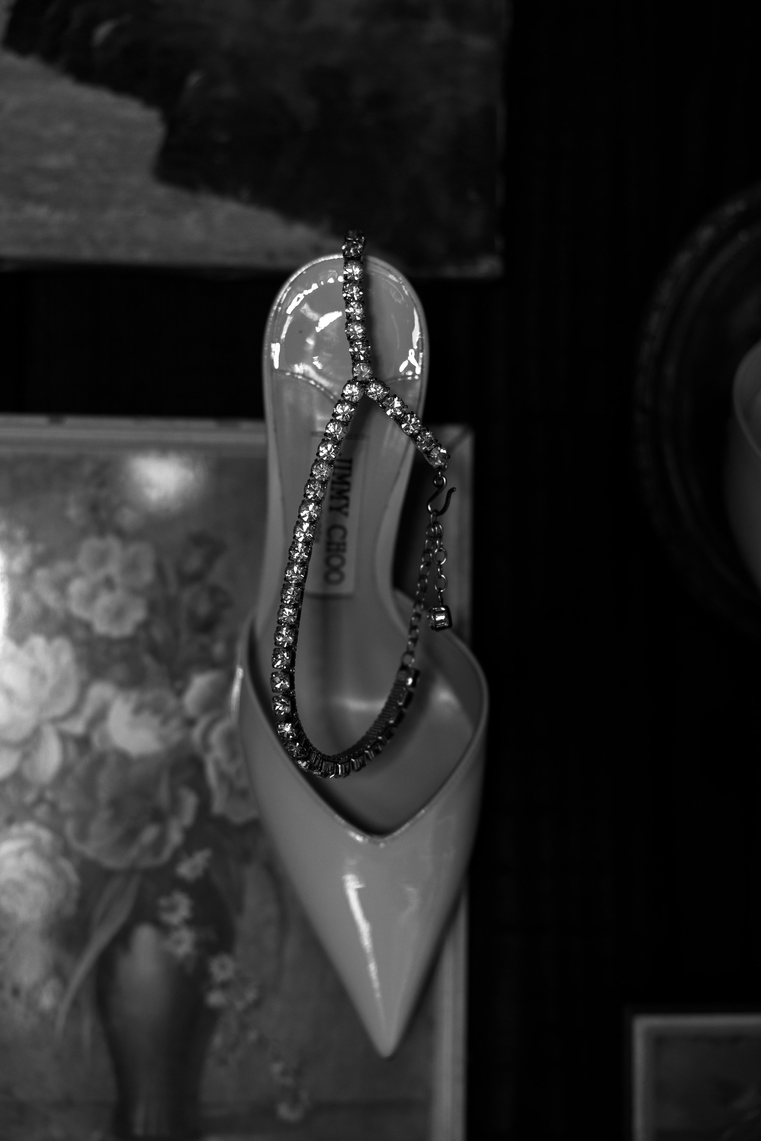 A pair of white high-heeled shoes with a rhinestone necklace draped over one shoe, set in front of a blurred floral painting and other objects. Wedding accessories. wedding photography. Editorial style photography. Gold Coast Australia.