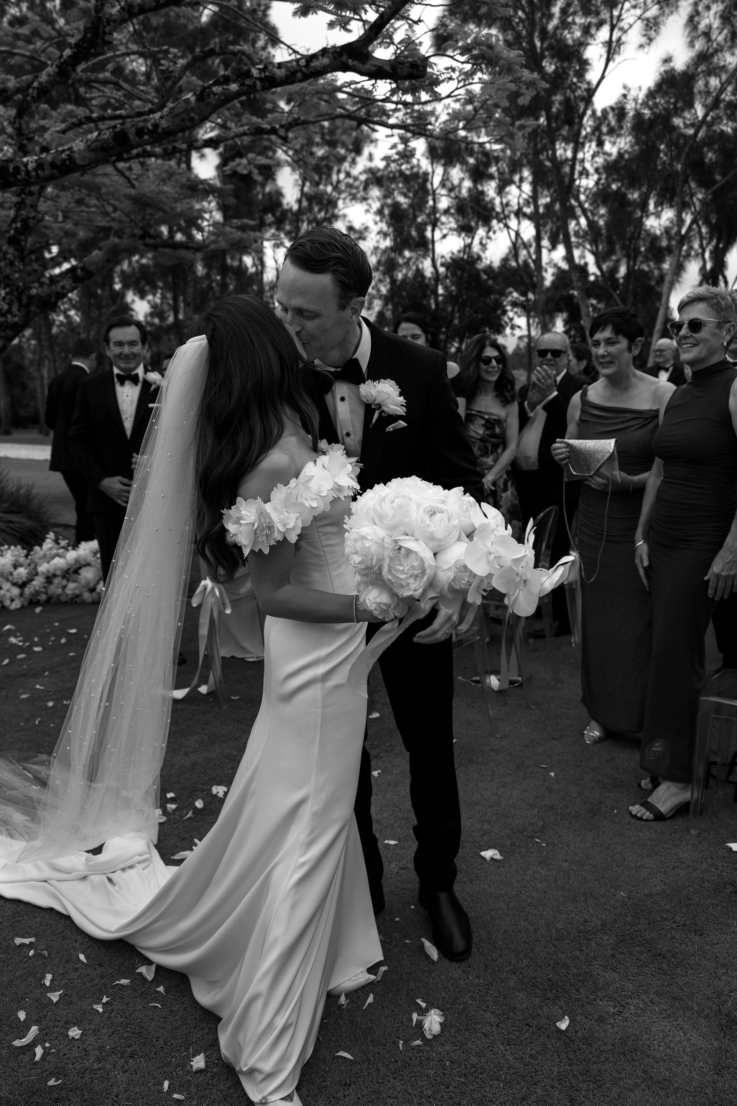 Black and white photo of a wedding ceremony showing a bride and groom sharing a kiss, surrounded by seated guests outdoors. The bride wears a gown and holding a bouquet of flowers. The groom is in a tuxedo. Guests are smiling and clapping.