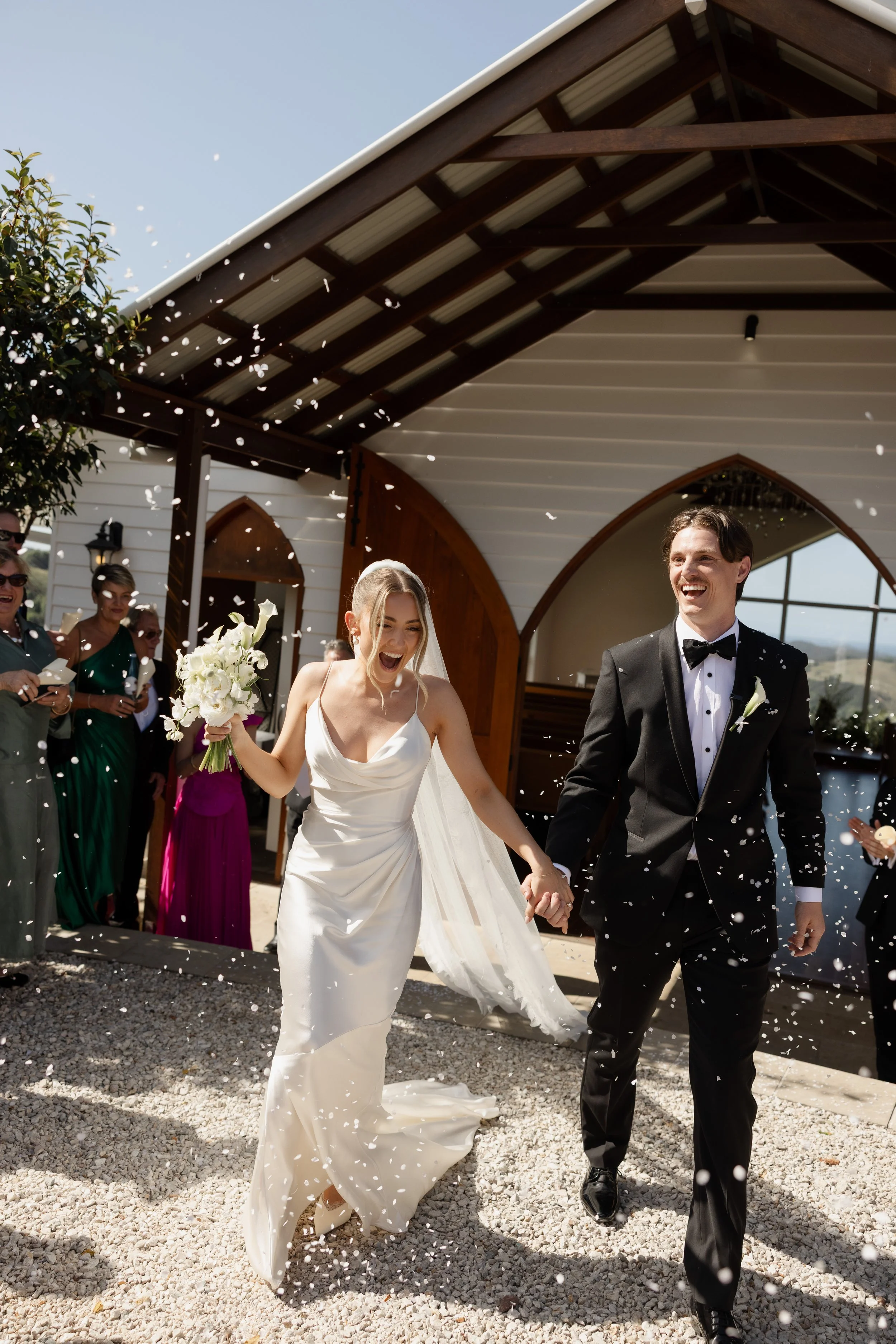 Four women in white dresses at a wedding, with three smiling and holding wine glasses, and one facing away holding a bouquet, standing outdoors near a pool with a background of green trees and red flowers. Gold Coast, Editorial Wedding Photography.