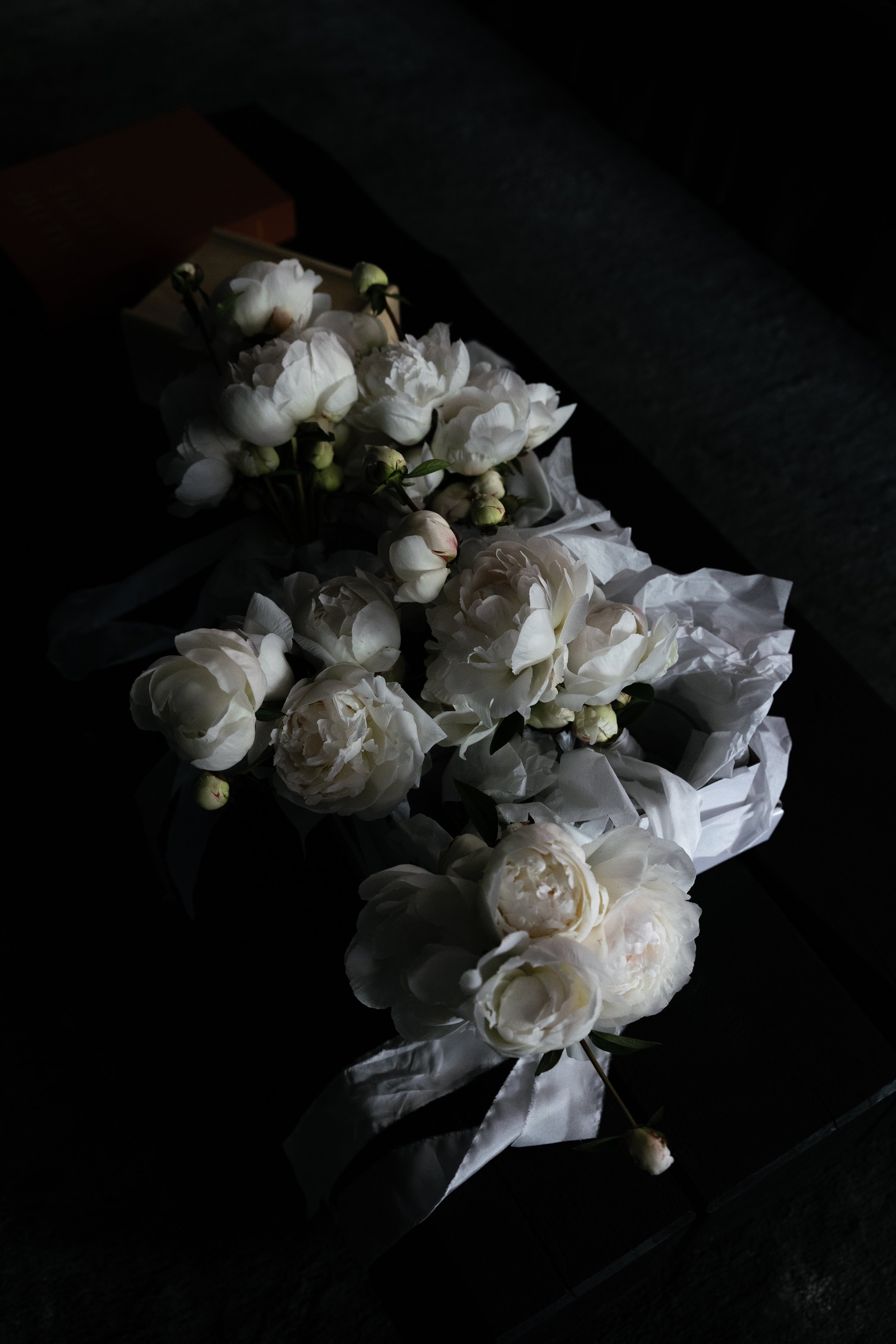 White peony flowers and small buds wrapped in white paper and ribbon, resting on a dark surface. Wedding accessories, black and white photography. Gold Coast Australia