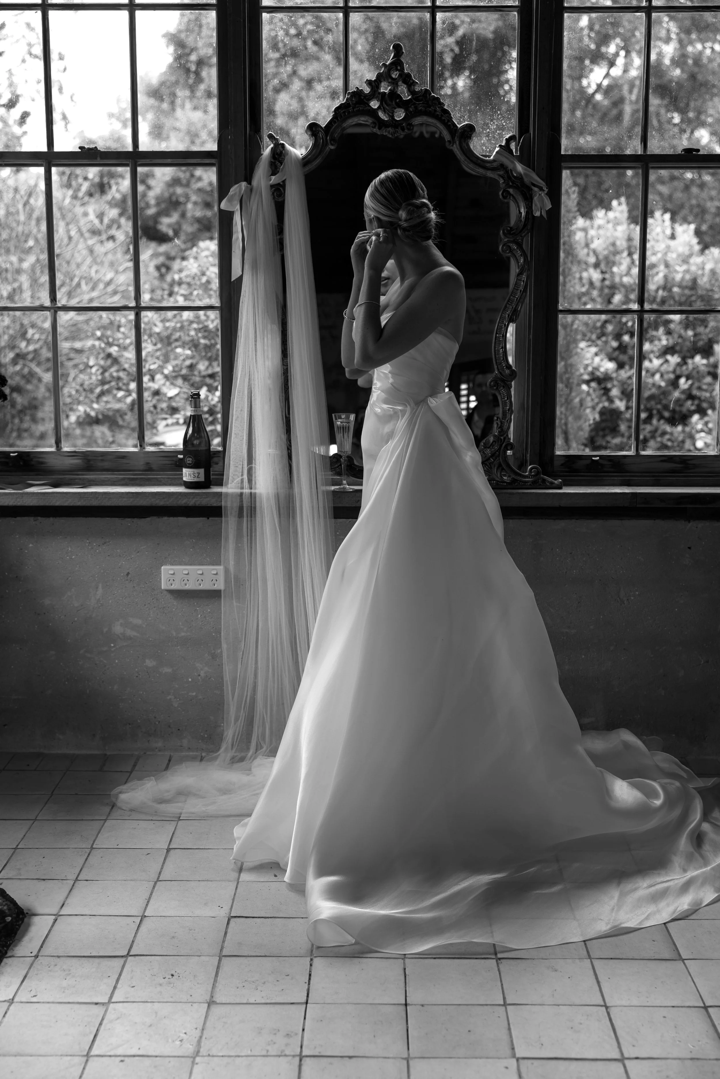 A bride in a white wedding dress standing in front of a large mirror, adjusting her earring. Byron Bay Australia Photography