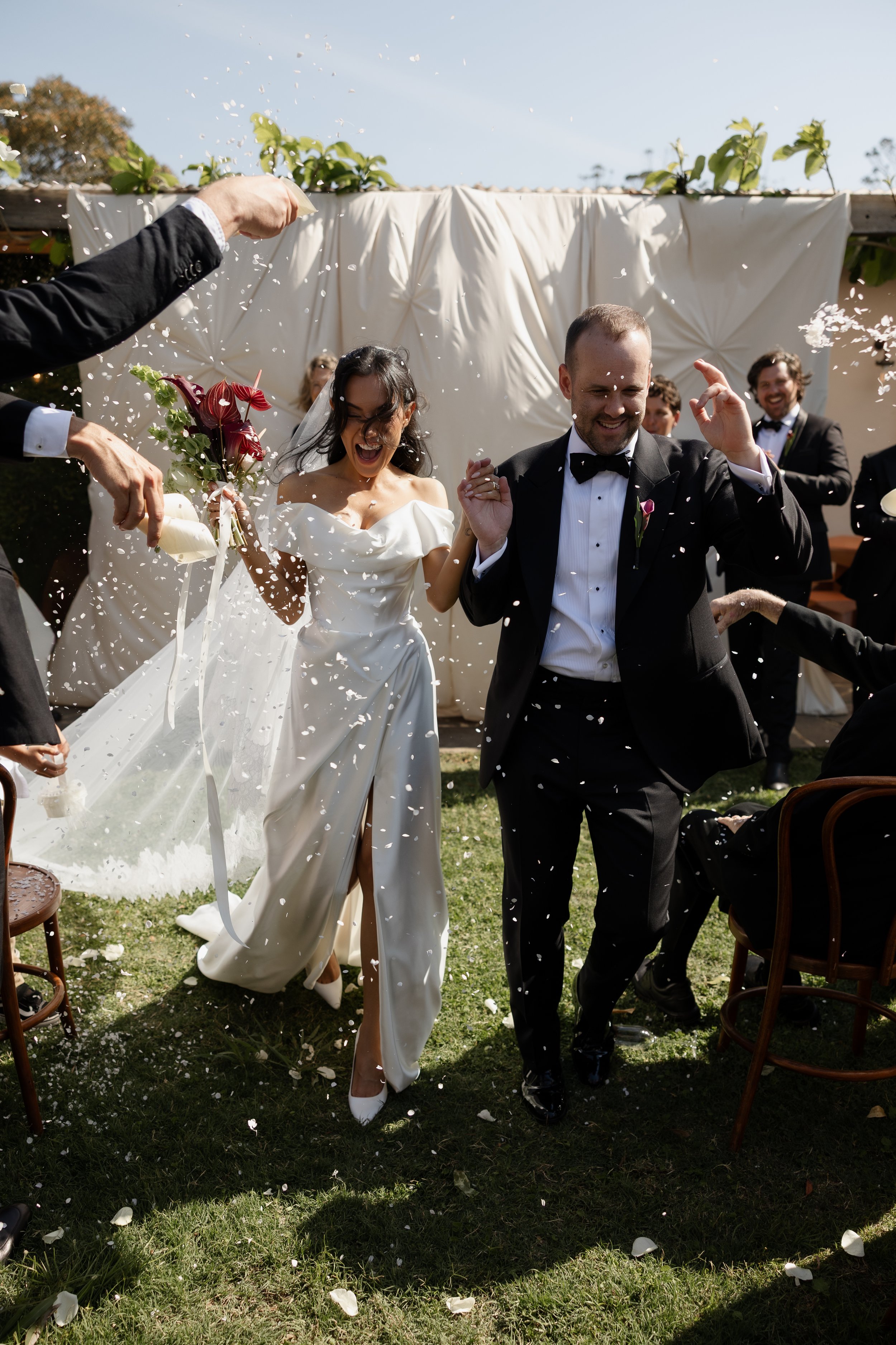 Four women in white dresses at a wedding, with three smiling and holding wine glasses, and one facing away holding a bouquet, standing outdoors near a pool with a background of green trees and red flowers. Gold Coast, Editorial Wedding Photography.