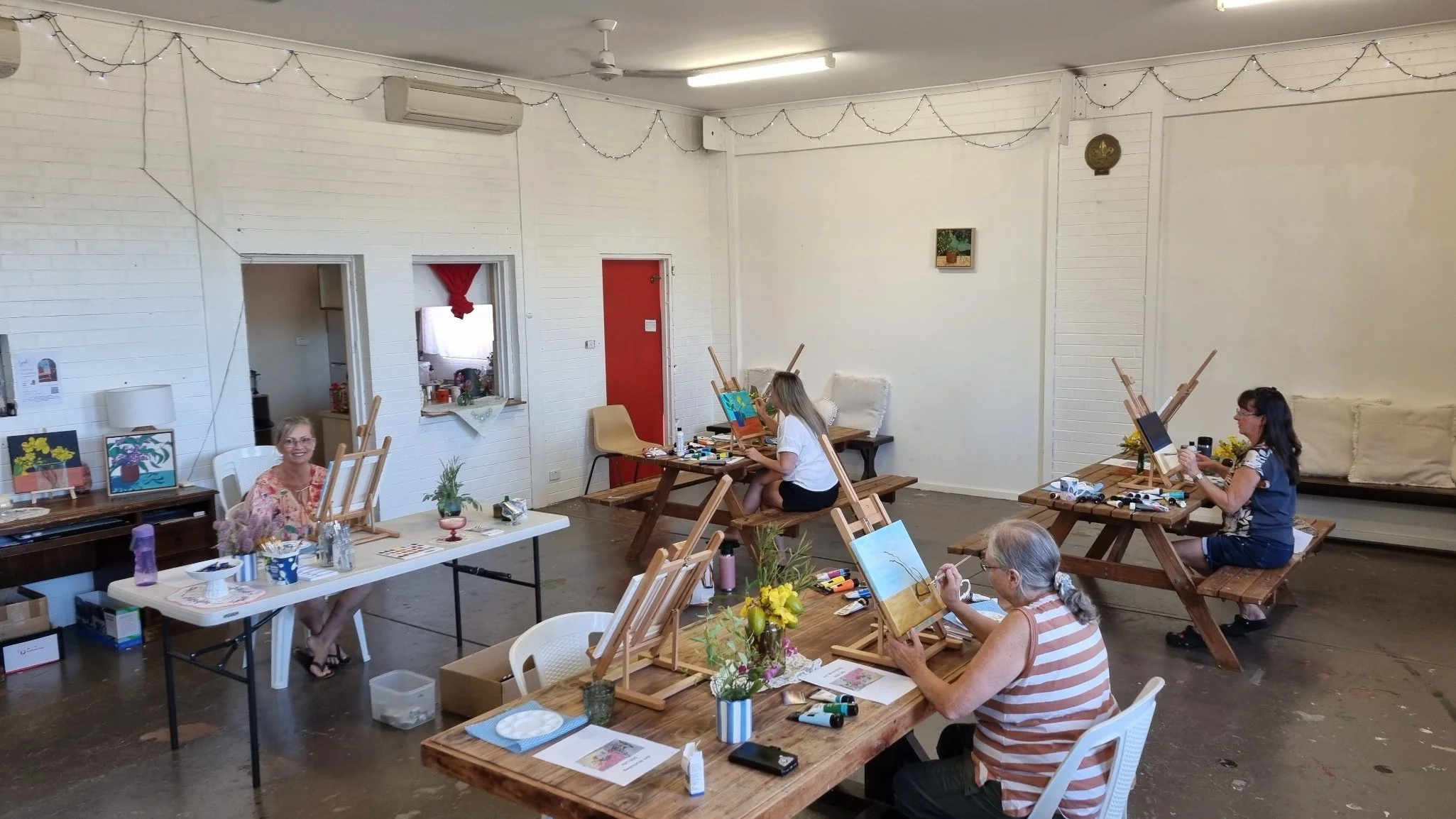 Indoor painting class with five women at tables, some painting on canvases, with easels, art supplies, and flower arrangements.