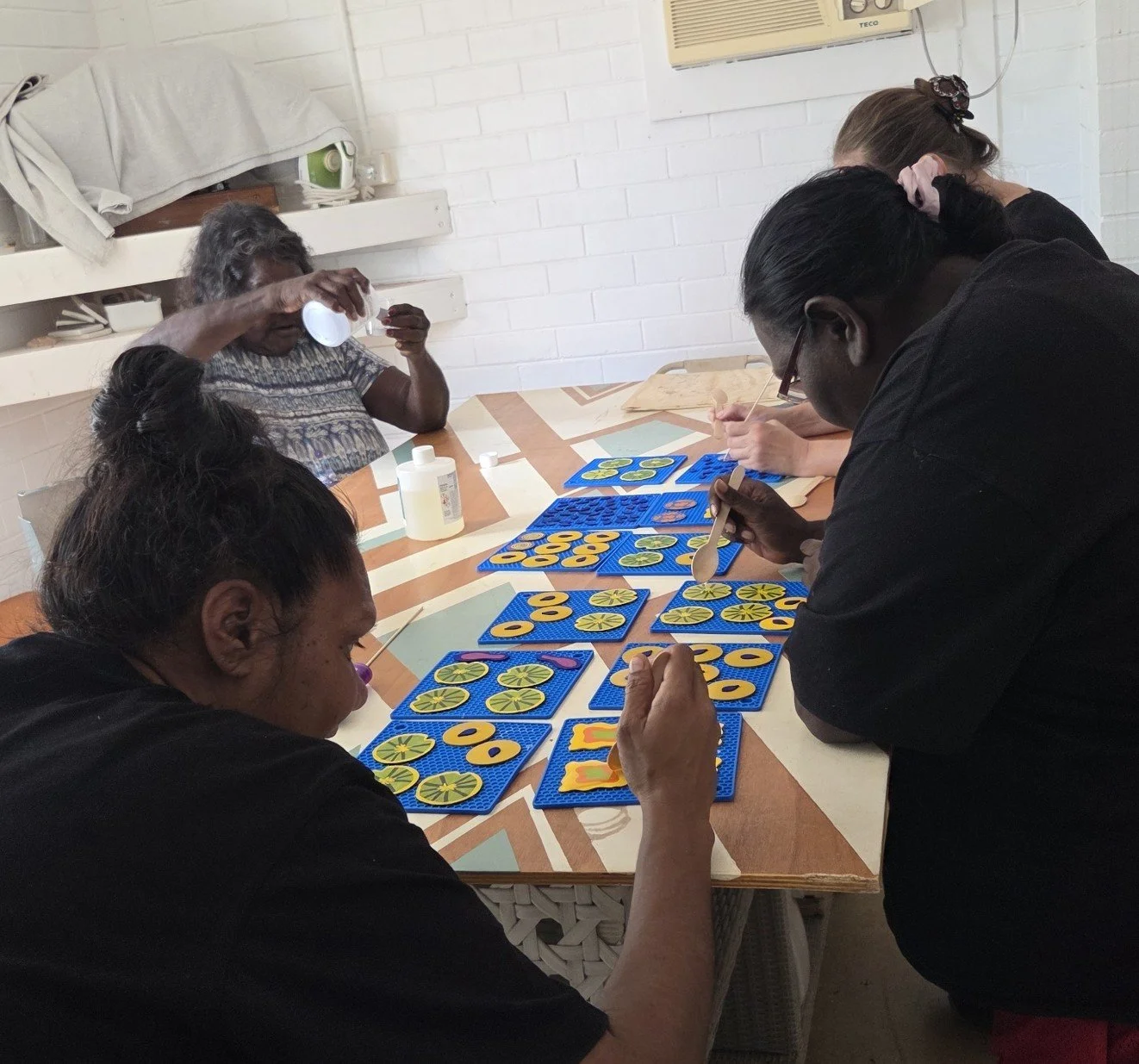 Four Australian Aboriginal women seated around a table engaging in a resin jewellery making activity with colorful circular and lemon-shaped pieces.