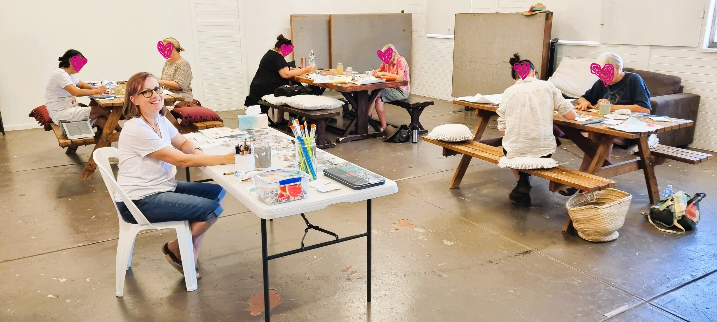 A group of women sitting around wooden tables in aworkshop, with one woman smiling at the camera. The tables are filled with supplies paper, and some women are engaged in creating projects while others are conversing. The room has a simple, industrial look with white brick walls and a concrete floor.