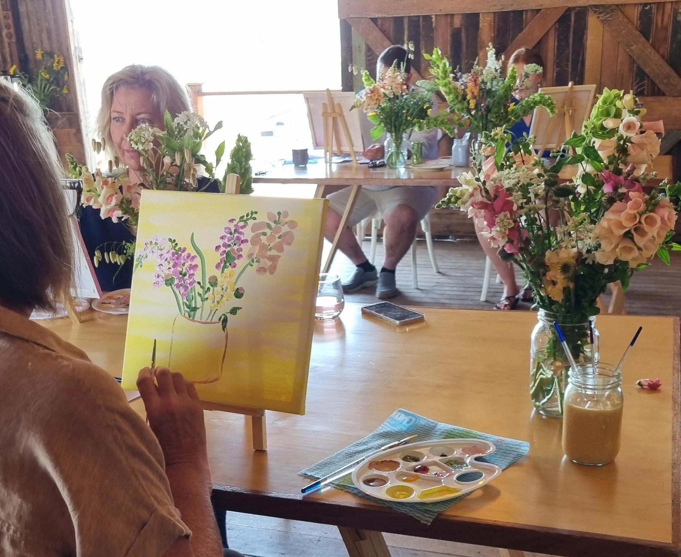 A woman painting a floral still life on a canvas at a table decorated with fresh flowers in a rustic room. The table holds a paint palette with various colors and brushes, along with a jar of water.