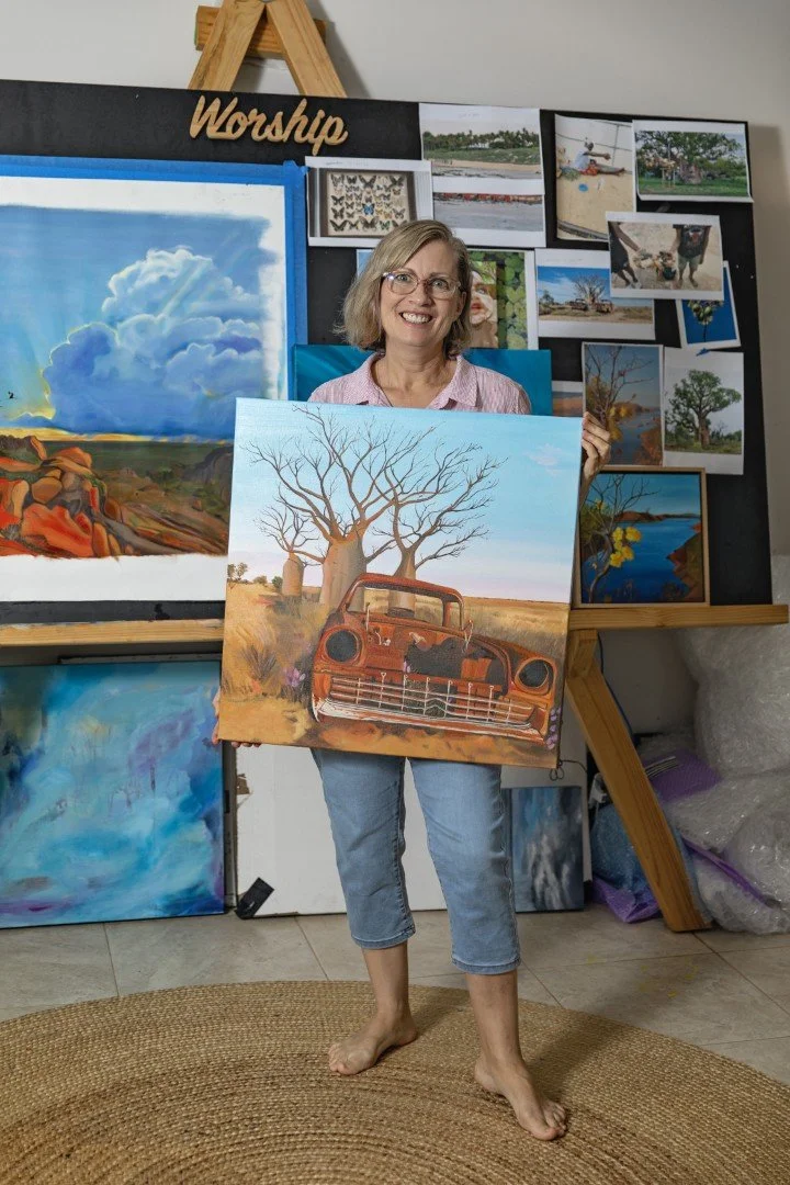 Woman in art studio smiling and holding a painting of a rusty old car in front of three boab trees.