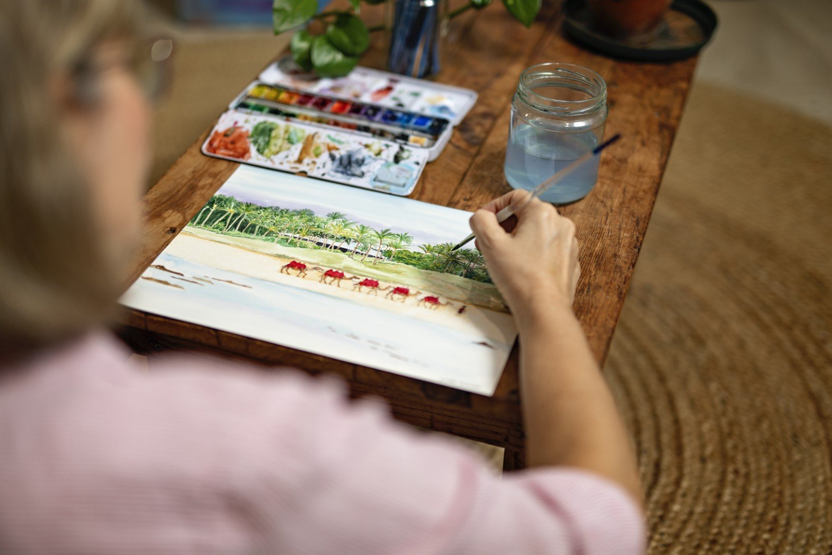 Female artist sitting at a low wooden table painting a small picture with watercolours.