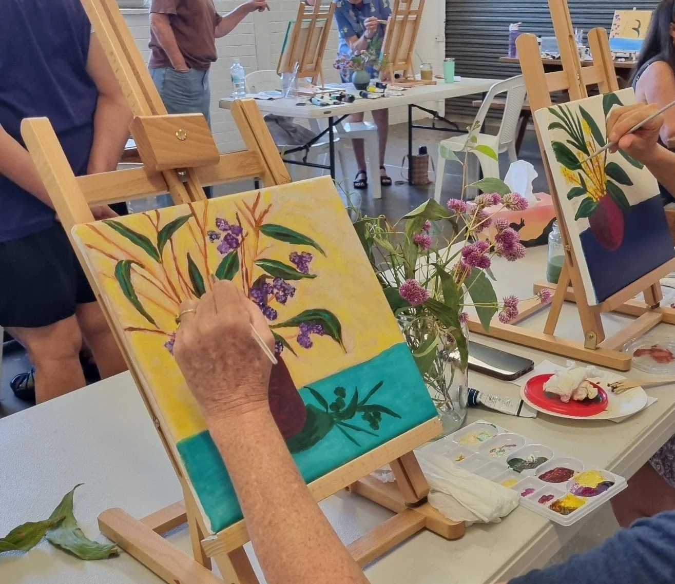 People painting floral still life on canvas at a group art class, with painting supplies, easels, and a vase with purple flowers on the table.