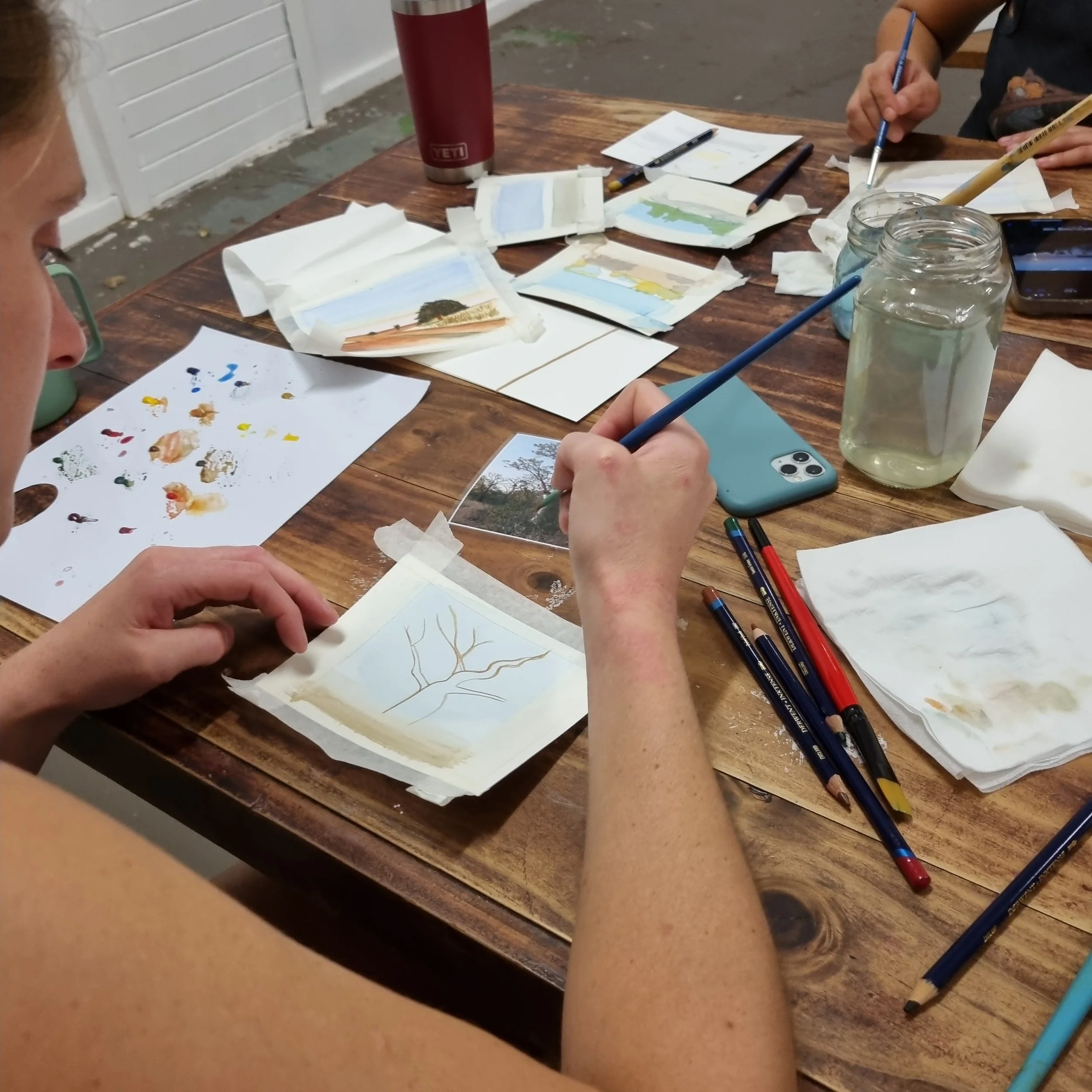 People painting miniature landscape scenes on small cards at a wooden table with watercolour paint supplies and water jars.