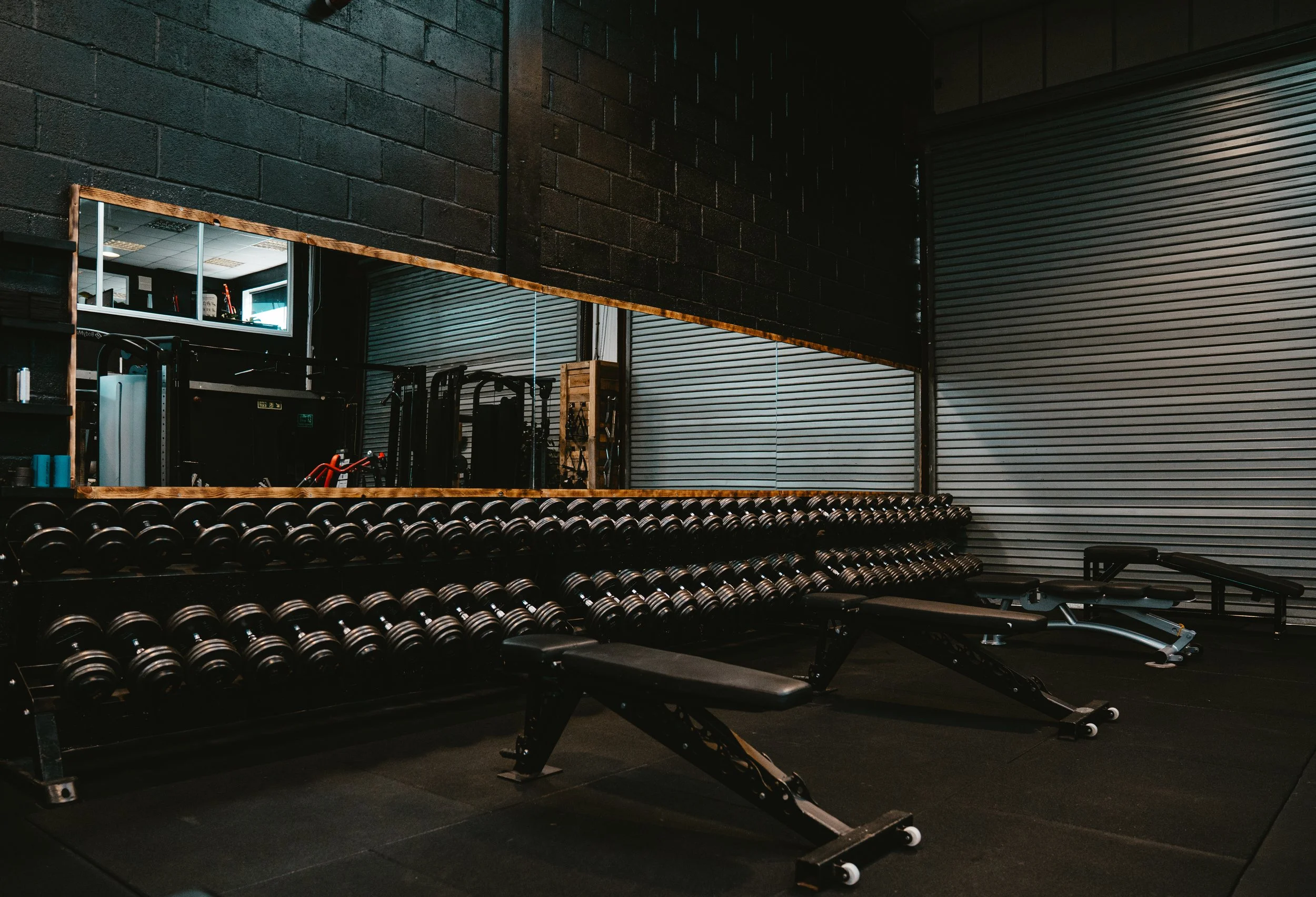 Interior of a gym with dumbbell racks, exercise benches, and a large mirror on the wall.