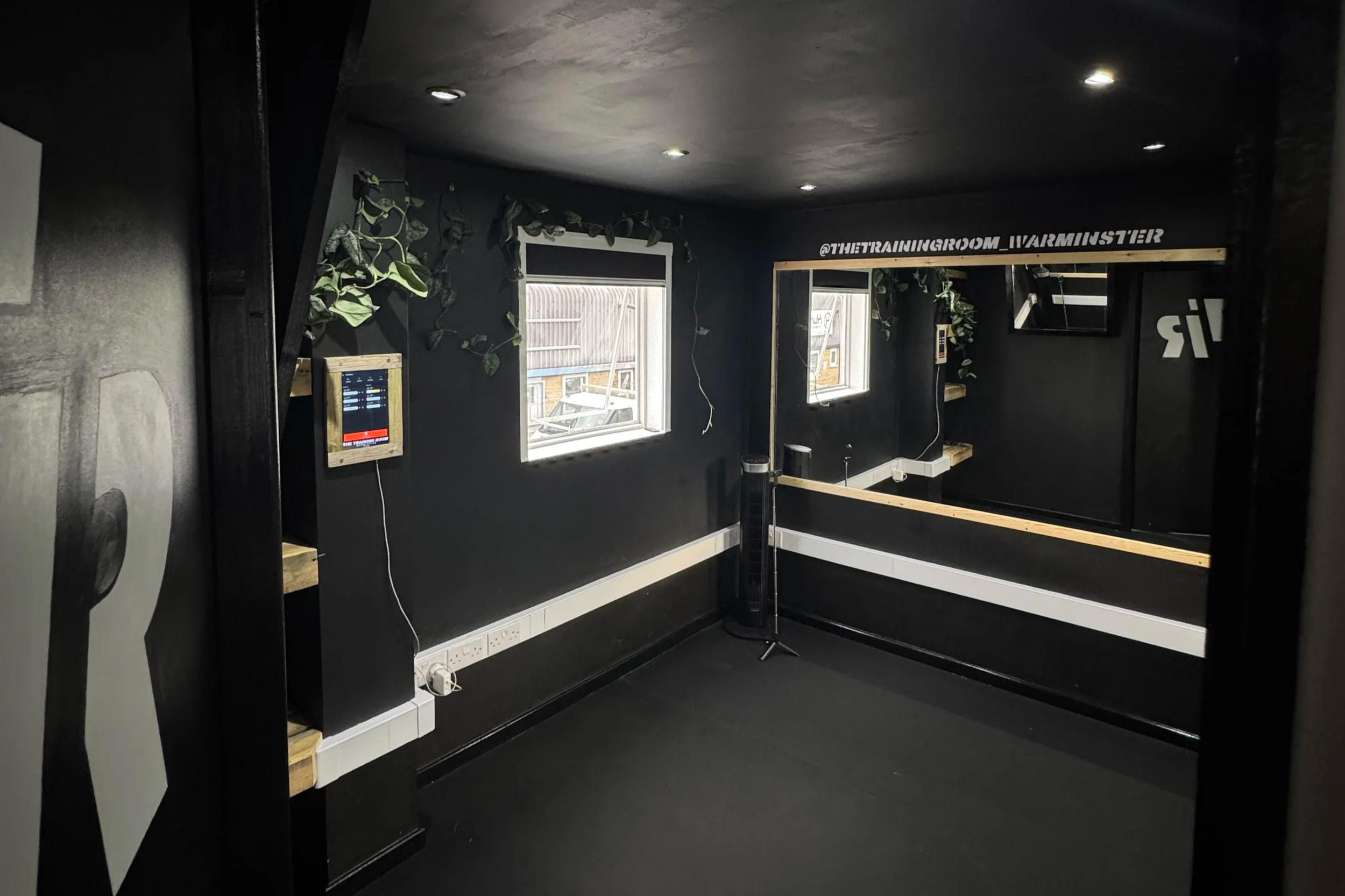 Black workout room with two windows, wooden shelves, a mirror, and a small black fan. Wall decor includes green leaves and a sign that reads '@THETRAININGROOM_WARMINSTER'.