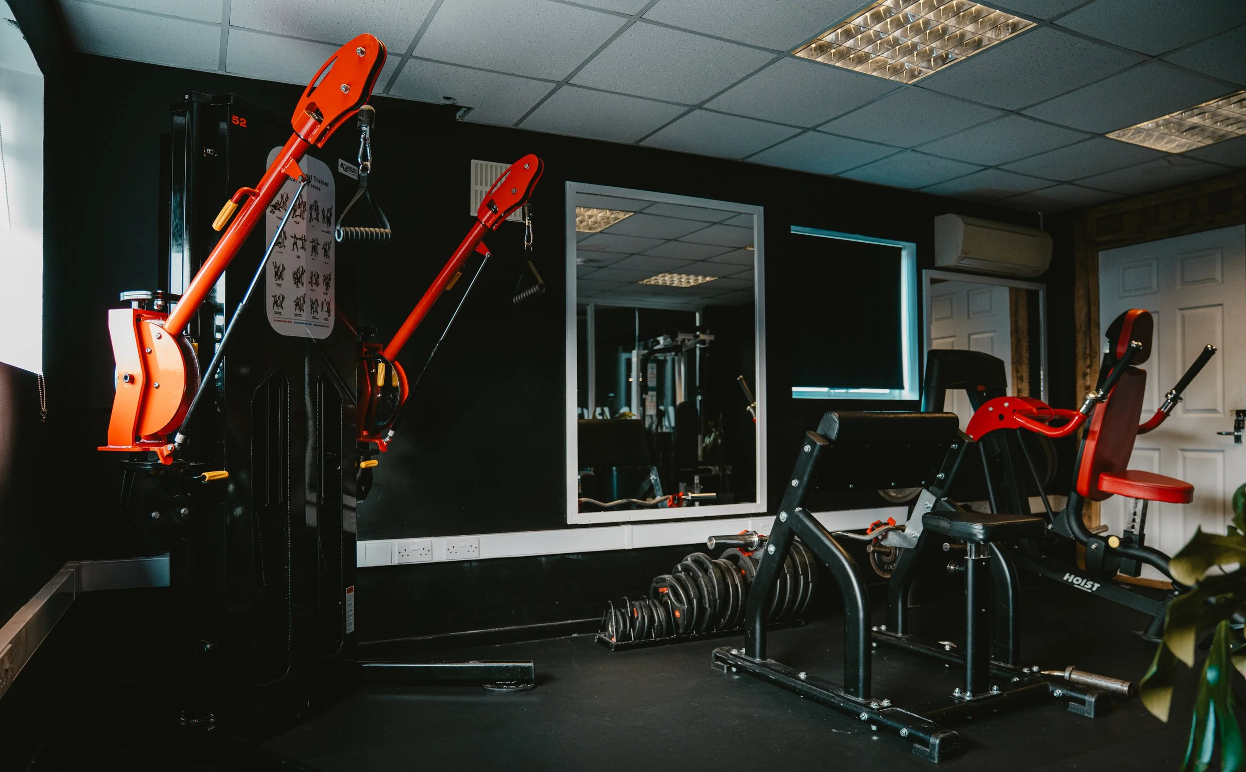 A small gym room with workout equipment including a leg press machine, an abdominal exercise machine, dumbbells, and a mirror on the back wall. There is a window with blue blinds and an air conditioning unit on the wall.