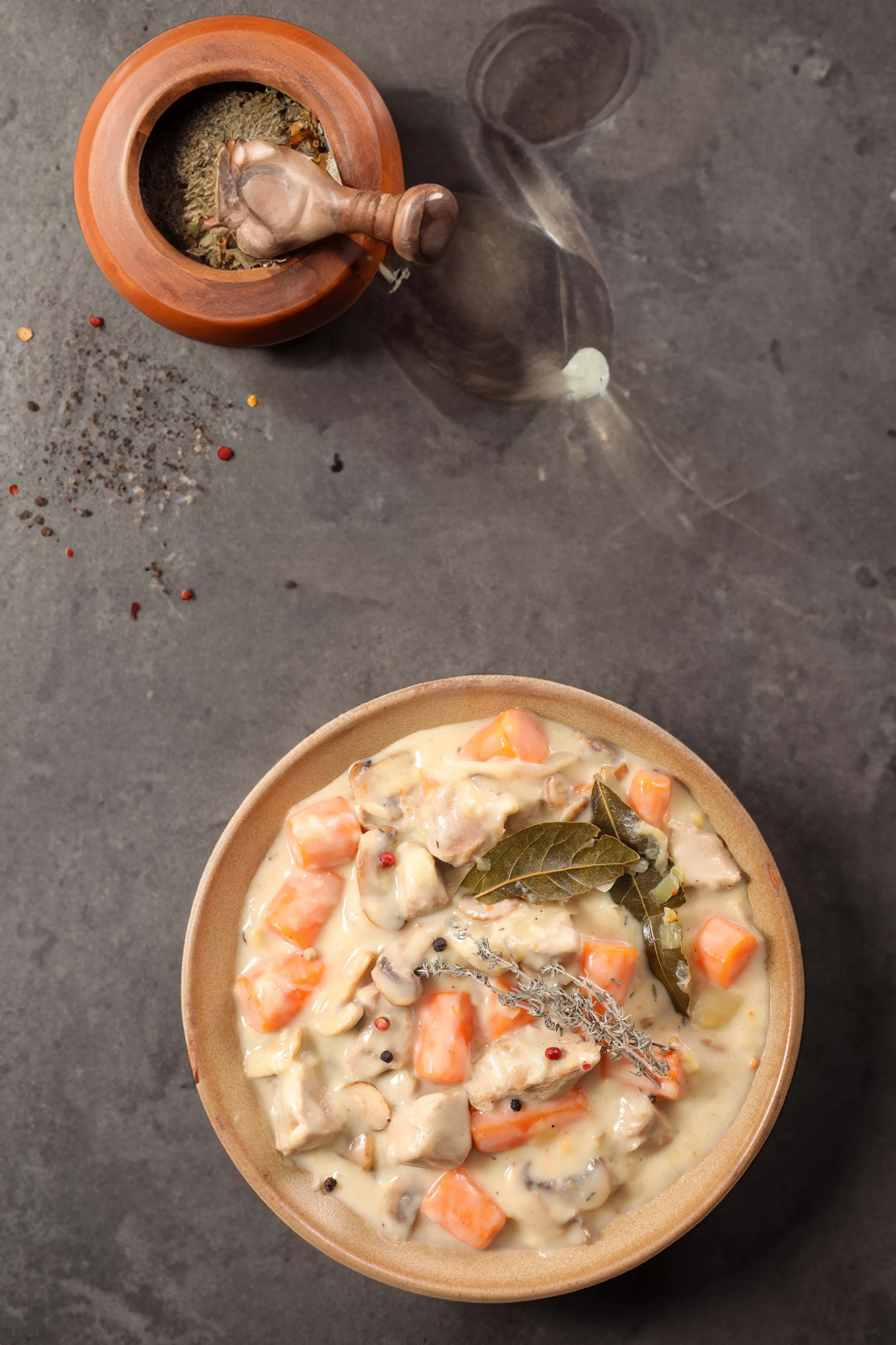 Een kom met warme stoofpot met stukjes wortel, vlees en champignons, gekruid met laurierblaadjes, op een zwarte tafel. Boven het bord ligt een lepel met witte saus en ernaast liggen peper en zout.