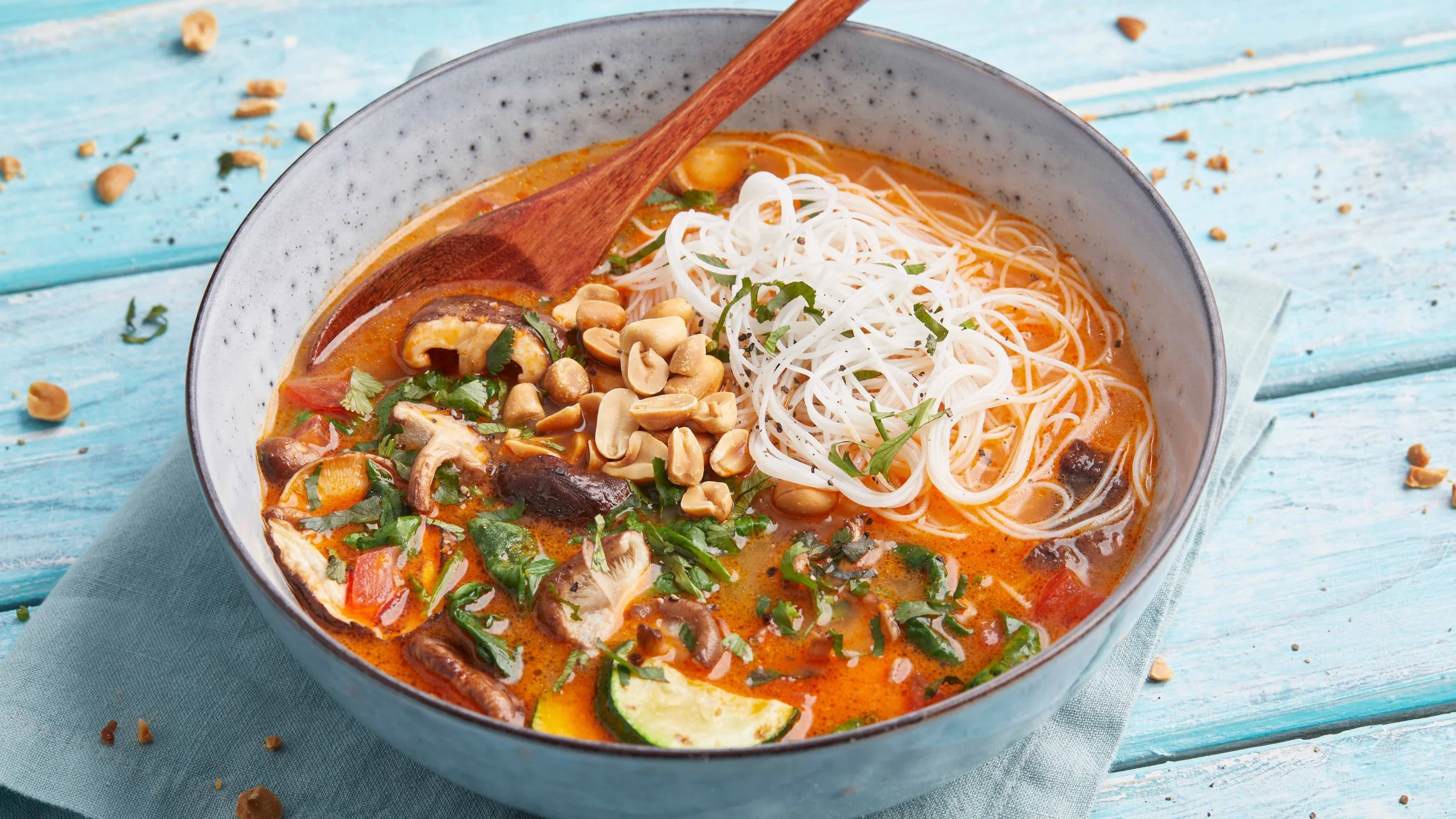 Een kom noedelsoep met groenten en pindas, geserveerd met een houten lepel op een blauwe houten tafel.