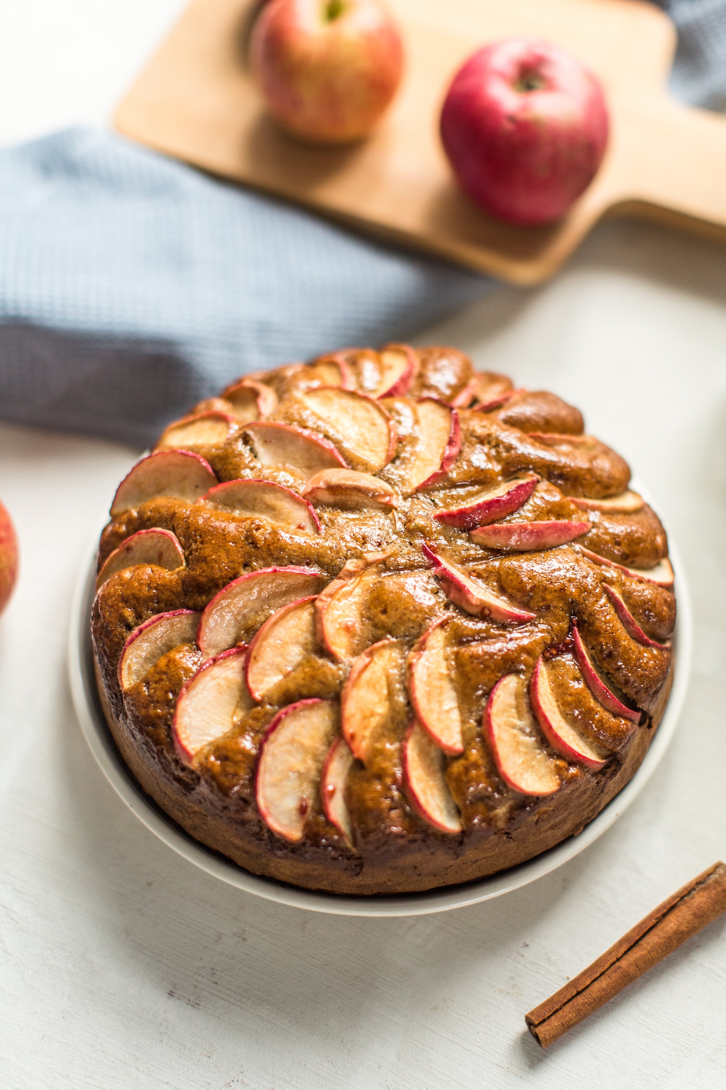 Appelkruimeltaart op een witte taartplateau met appels en kaneelstokje ernaast