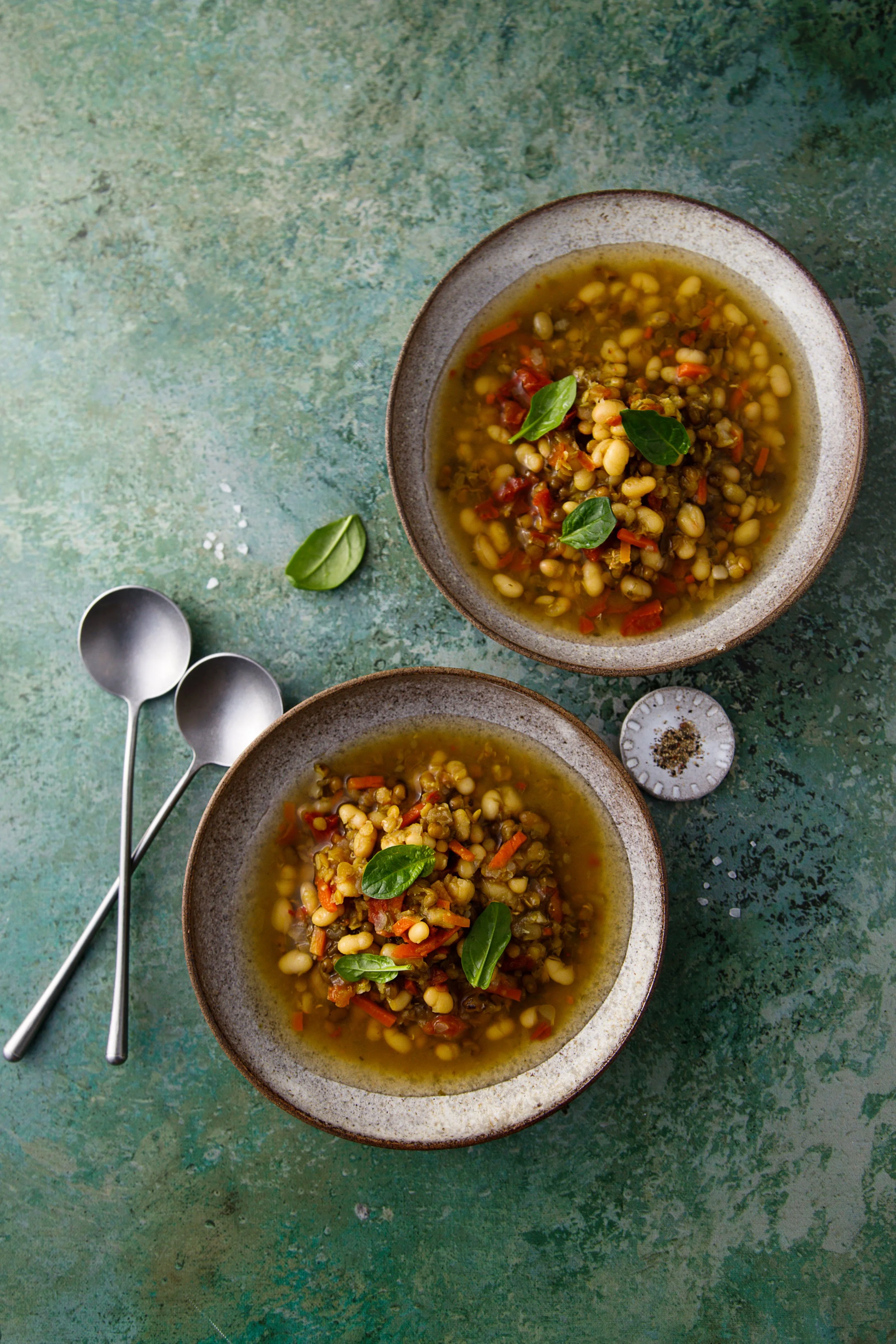 Twee kommen met linzensoep op een groene tafel, versierd met basilicumblaadjes, naast twee lepels en een potje met peper.