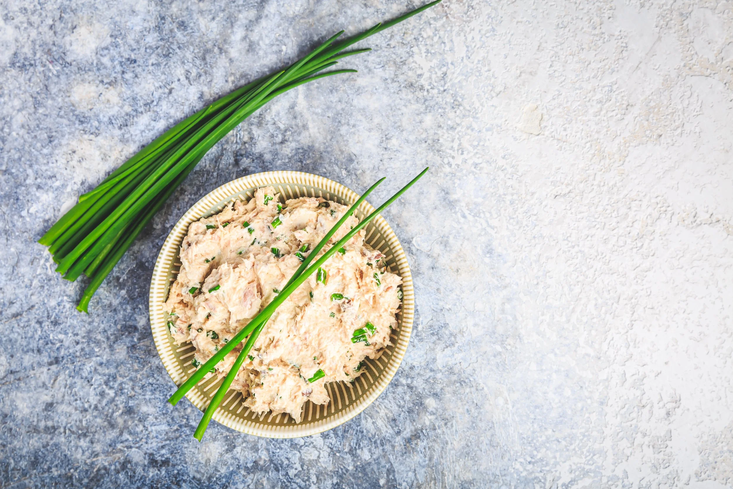 Kleine schaal met een tonijnsalade, naast een bosje groene uien op een blauwe en witte gestructureerde achtergrond.