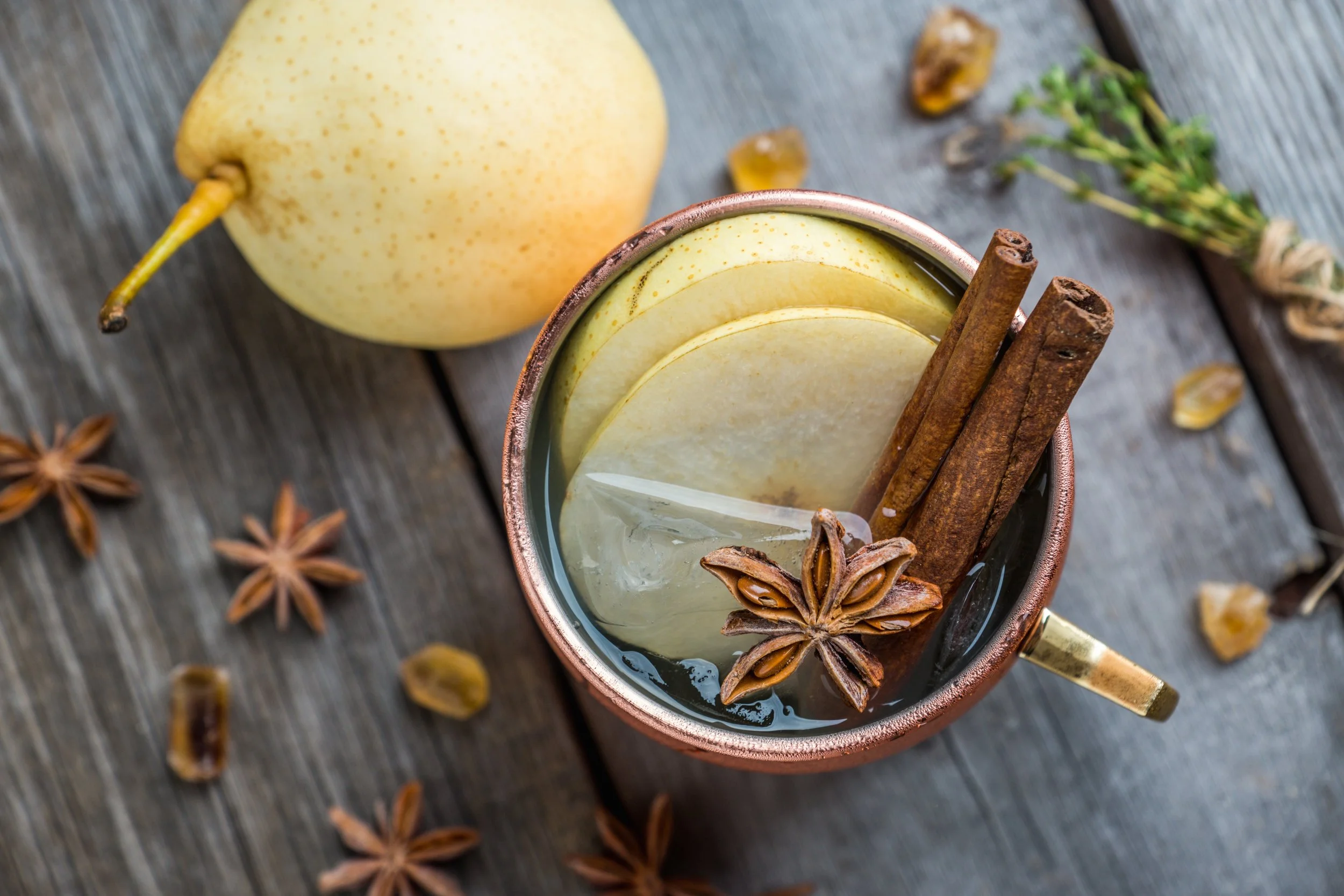 Een kopje met warme drank met schijfjes peer, kaneelstokjes, een steranijs en ijsblokjes, naast een hele peer en wat kruiden op een houten tafel