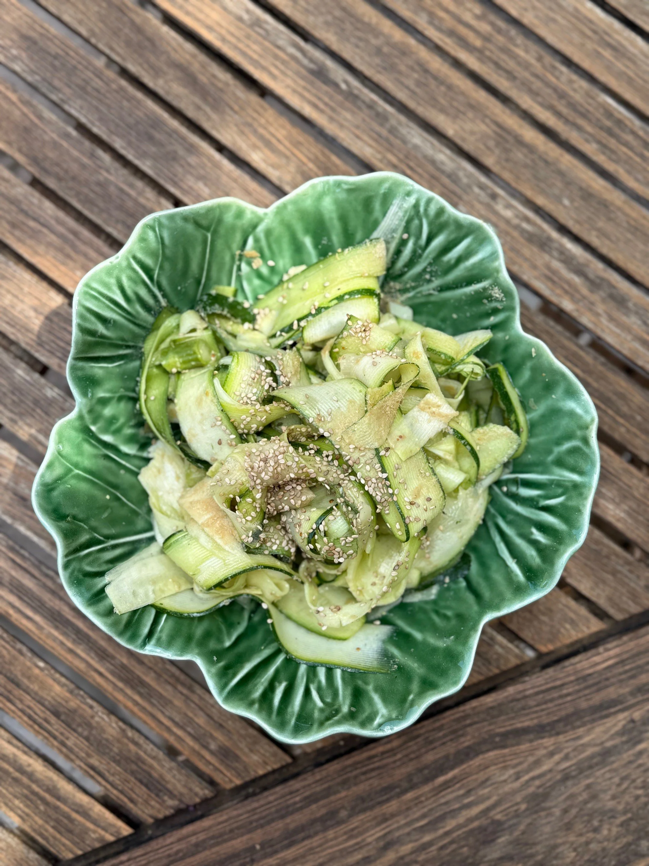 Kommet met dunne plakjes komkommer, bestrooid met sesamzaadjes, op een groene, bladvormige schaal op een houten tafel.
