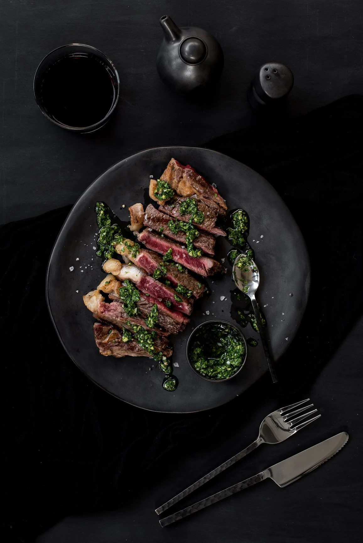 Gekookt stukje vlees met groene kruiden en saus op een zwart bord, naast een mes en vork, met een glas wijn en zout- en pepervaatjes op een zwarte tafel.