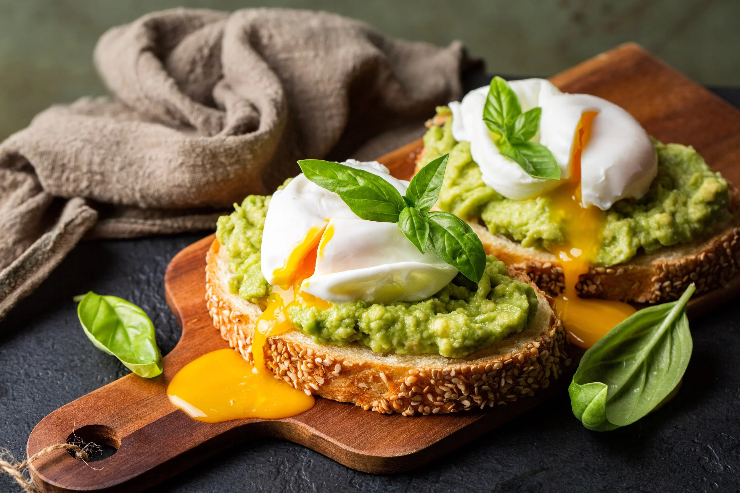 Geroosterde sesamzaadgebakken brood met avocado, gepocheerd ei en verse basilicumblaadjes, geserveerd op een houten snijplank.