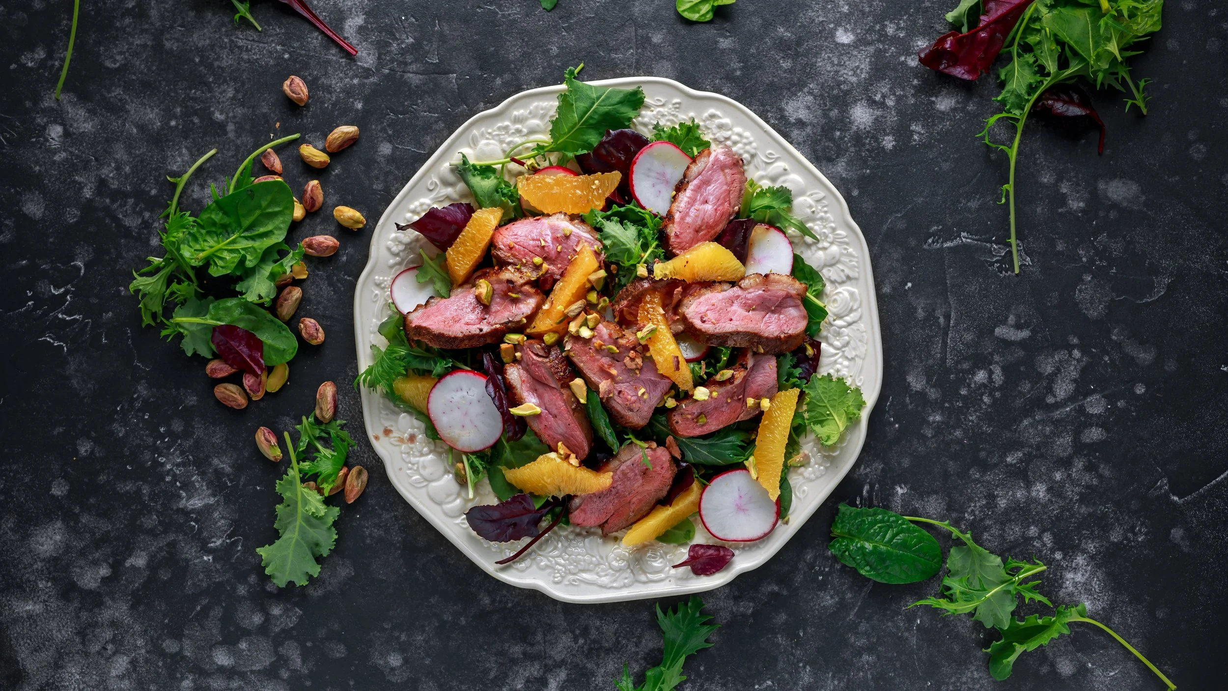 Salade met gegrilde steak, sinaasappelpartjes, radijs en gemengde greens op een wit bord.