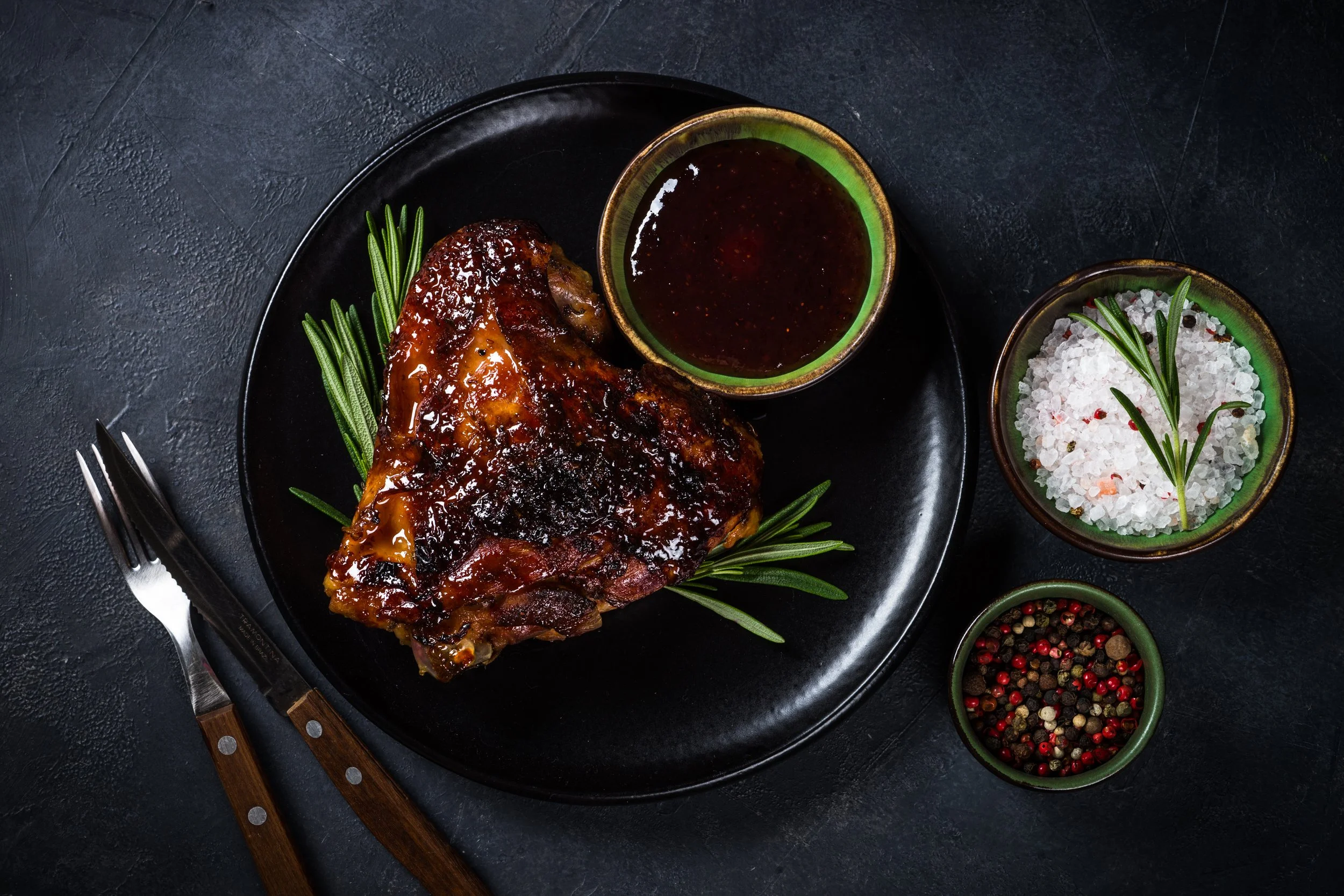 Geroosterd stuk vlees met groene kruiden, geserveerd met saus, zout, en peper in kleine kommen op een zwarte tafel.