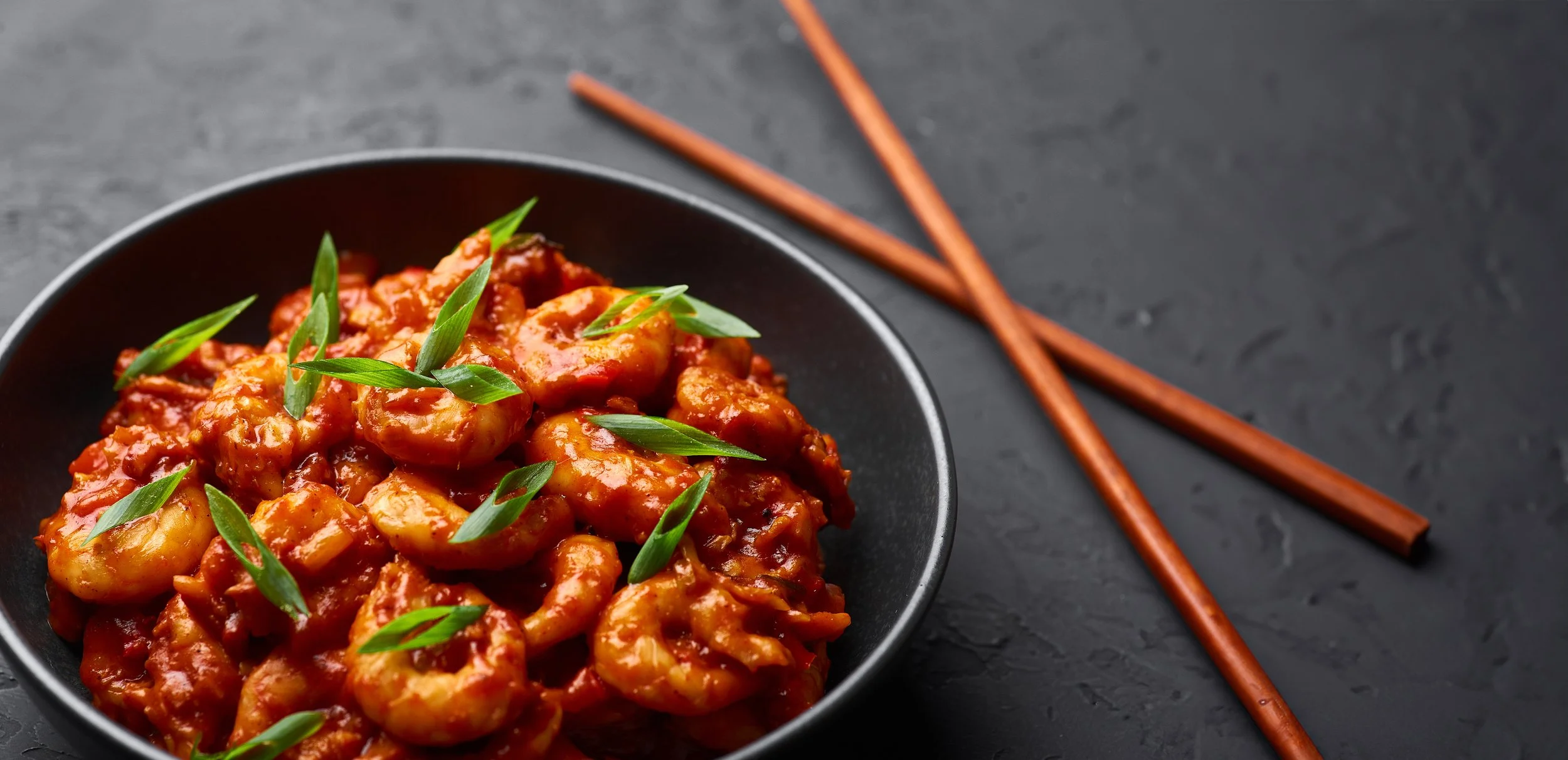 Bowl met garnalen in saus, garnalen zijn gekookt en bedekt met een rode saus, gegarneerd met groene uien, op een donkere tafel met een paar rode eetstokjes ernaast.