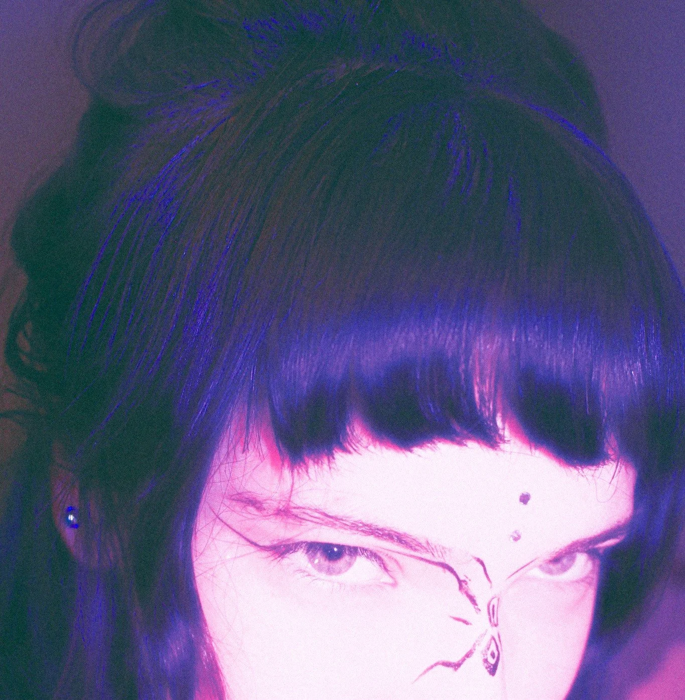 A close-up of a person's face with dark hair, light-colored eyes, and a butterfly drawing on their forehead.