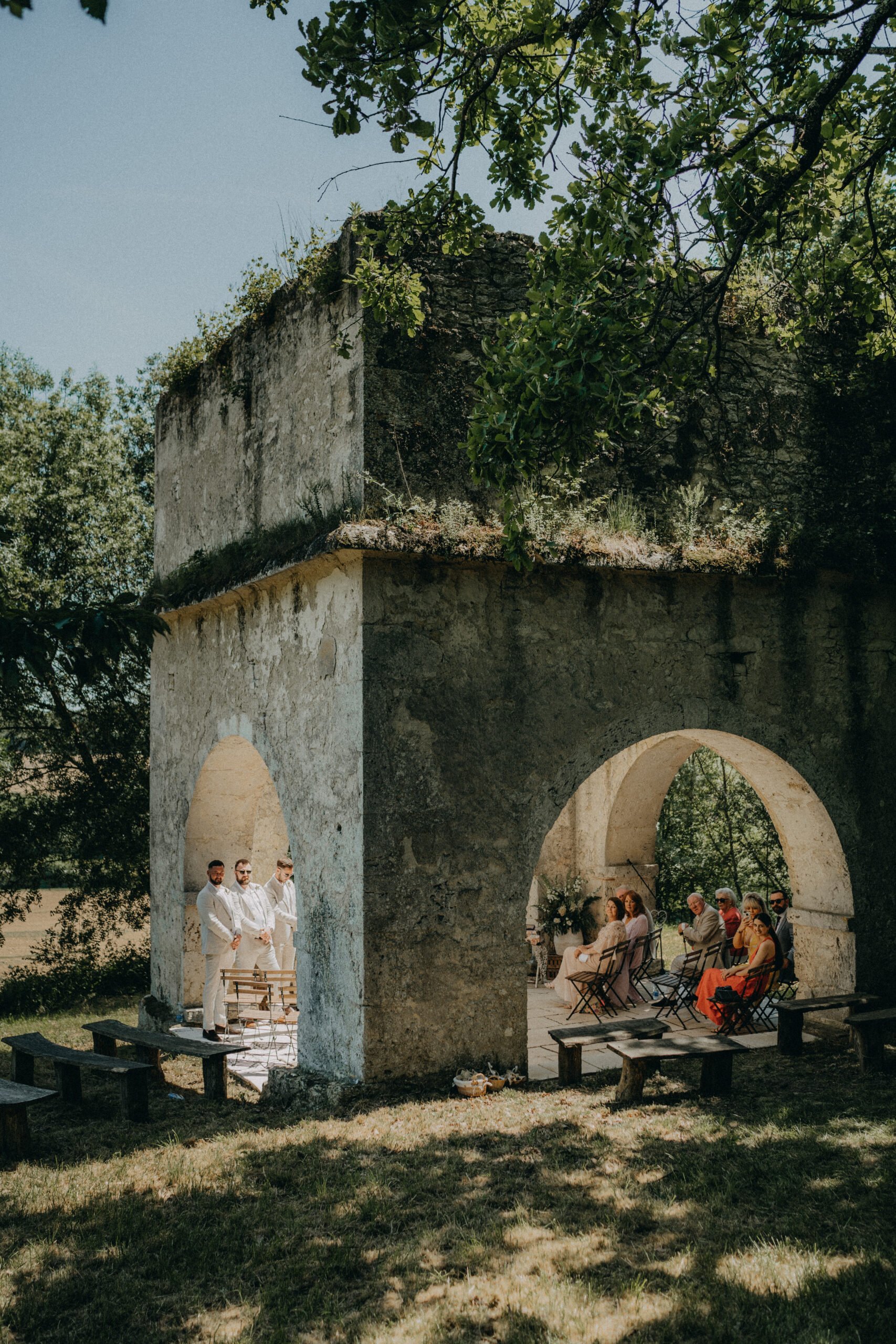 Outdoor wedding ceremony with guests seated under an old stone structure with arches, surrounded by trees.