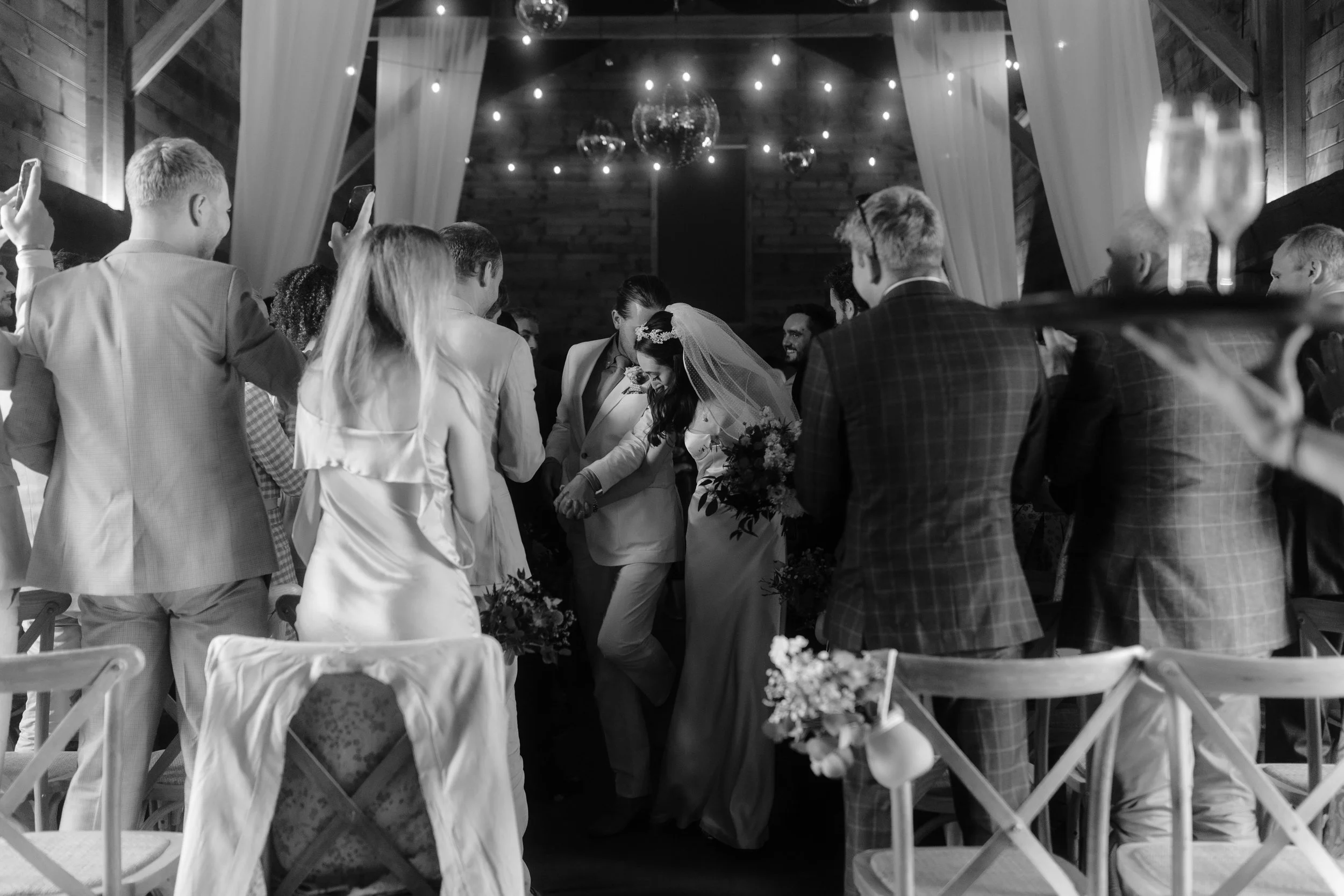 Black and white photo of a wedding ceremony with a bride and groom walking down the aisle, surrounded by guests clapping and taking photos. The setting is decorated with curtains and string lights.