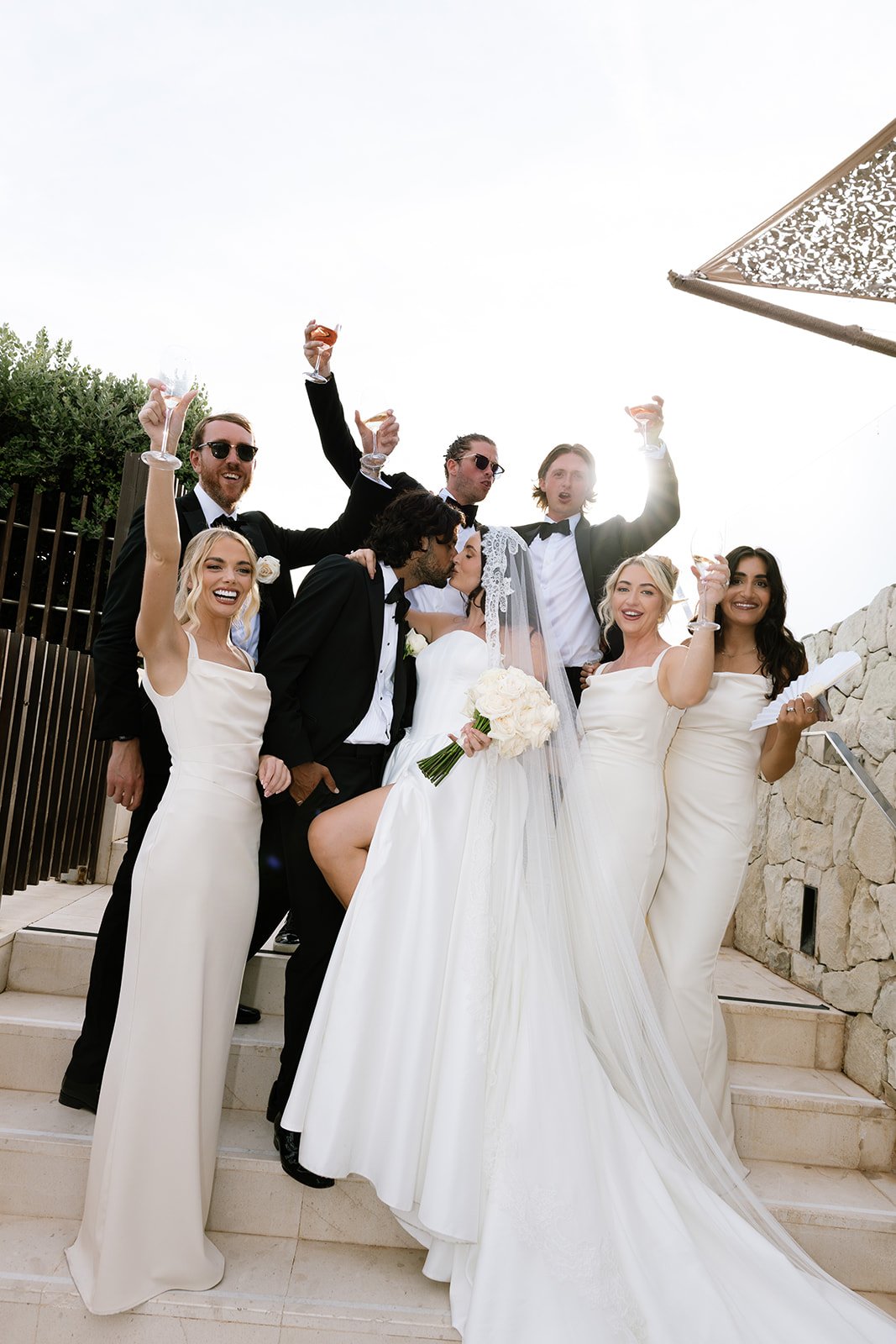 A sunlit photo of a kissing couple surrounded by their bridal party on the steps of their wedding venue.