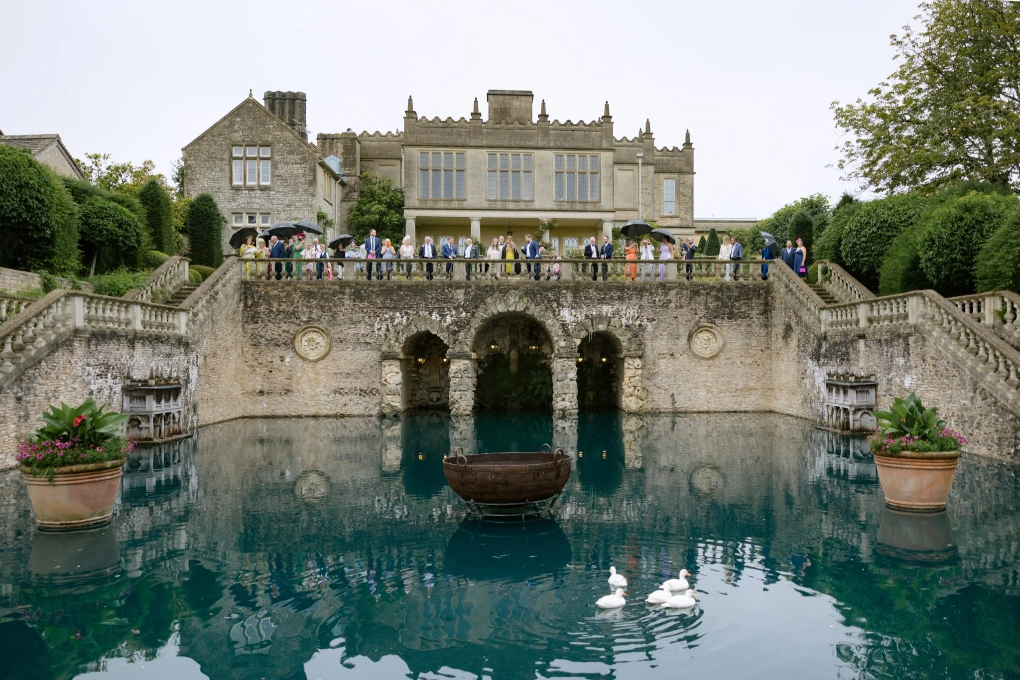 Guests gathered along the stone balcony overlooking the iconic turquoise water terrace at Euridge Manor in the Cotswolds, planned by Stacey Koks Weddings.