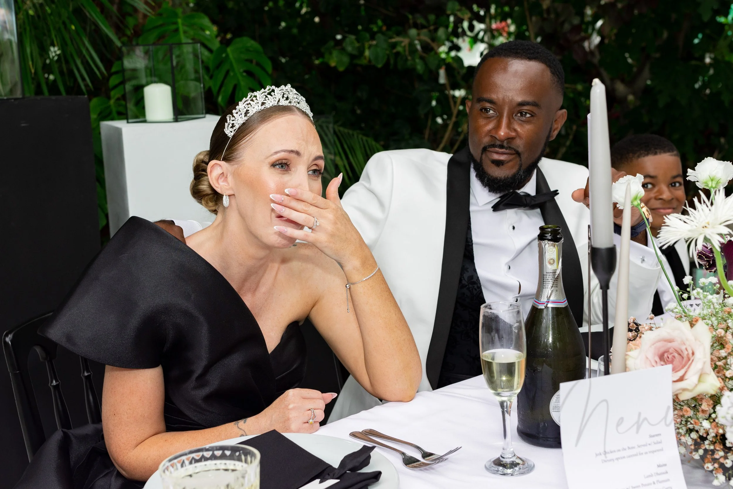 A couple at a wedding reception. The woman is wearing a black dress and a tiara, covering her mouth with her hand, appearing emotional. The man is wearing a white tuxedo with a black bow tie, sitting beside her. On the table, there are champagne glas