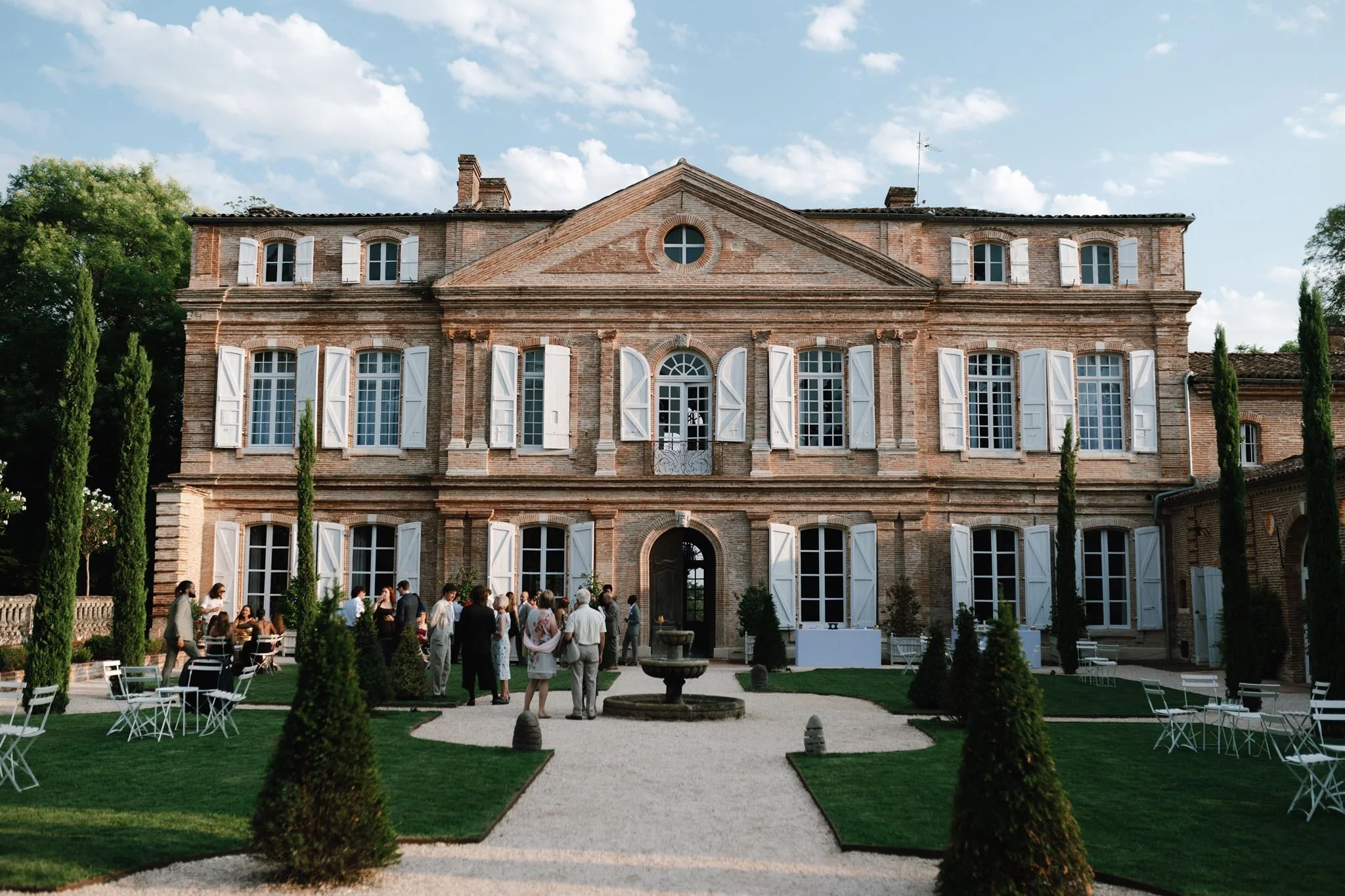 Guests enjoying a luxury outdoor drinks reception in the manicured gardens of an authentic brick chateau in France, produced by Stacey Koks Weddings.